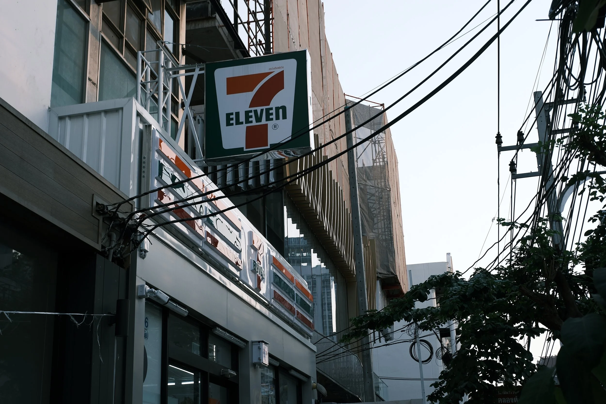 A street scene with a 7-Eleven convenience store sign on an urban building, multiple electrical wires overhead, and trees in the foreground.