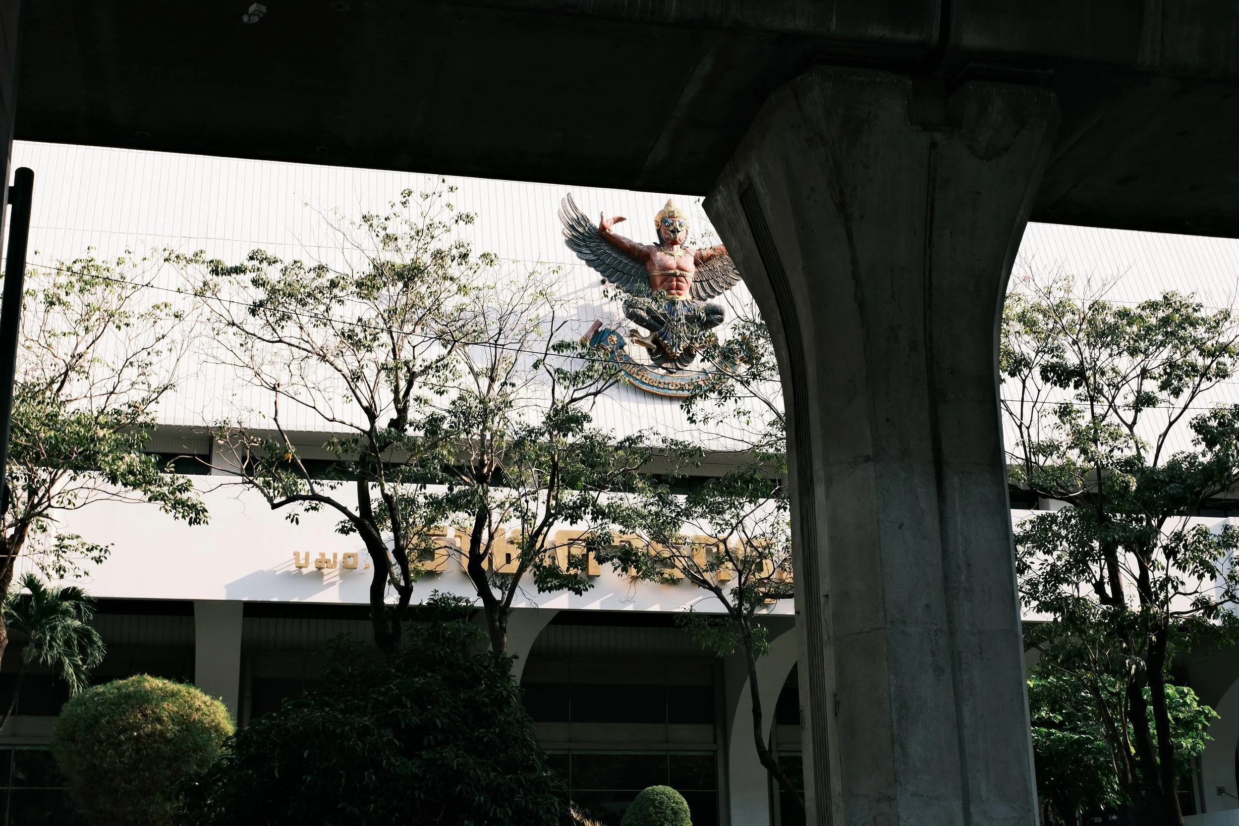 A large, colorful sculpture of a mythological creature with a humanoid upper body, wings, and a snake-like lower body is mounted on the roof of a building, seen through trees and under an overpass.