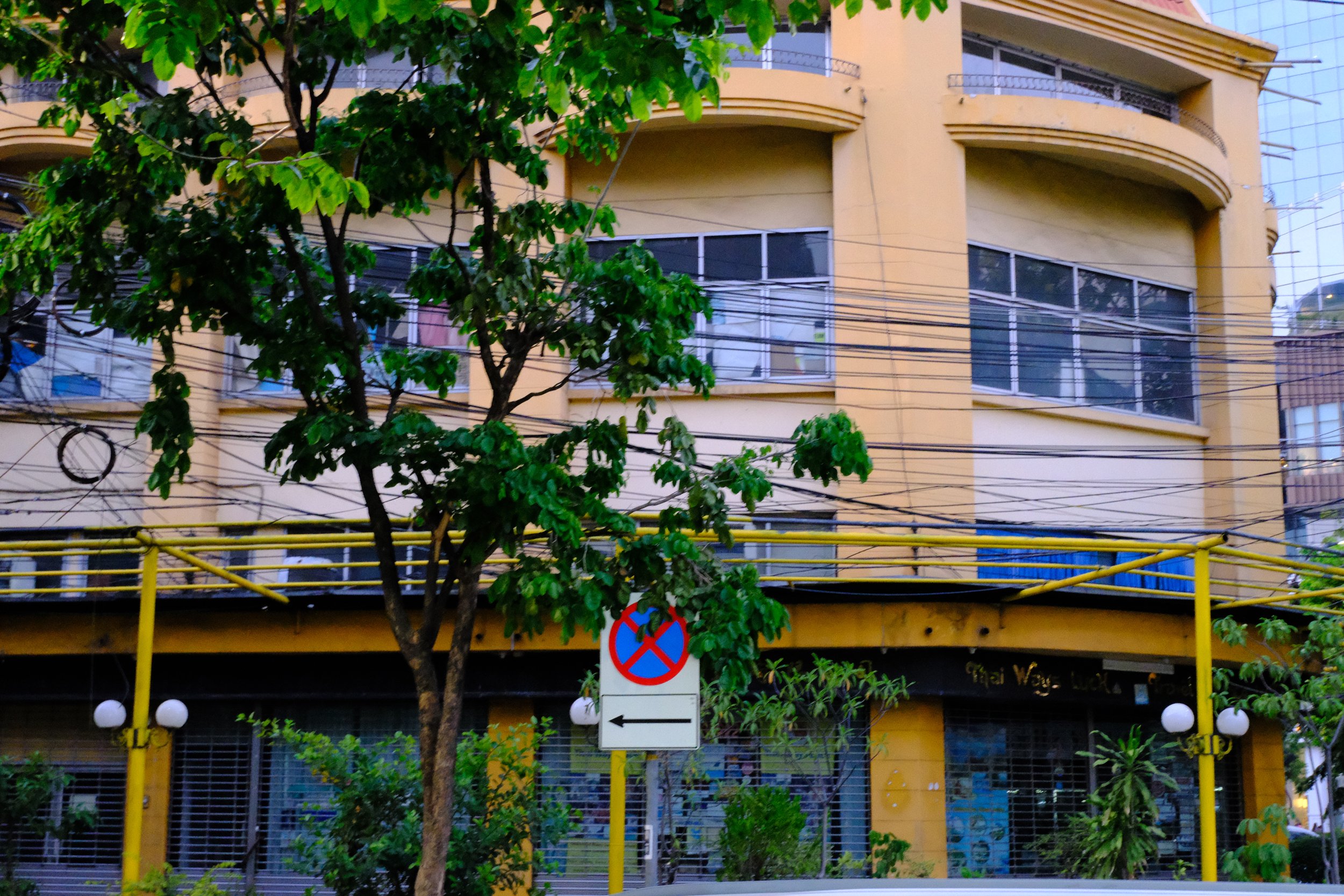 A yellow building with large windows, a tree in front, numerous electrical wires overhead, and a street sign with a left arrow and a no parking symbol.