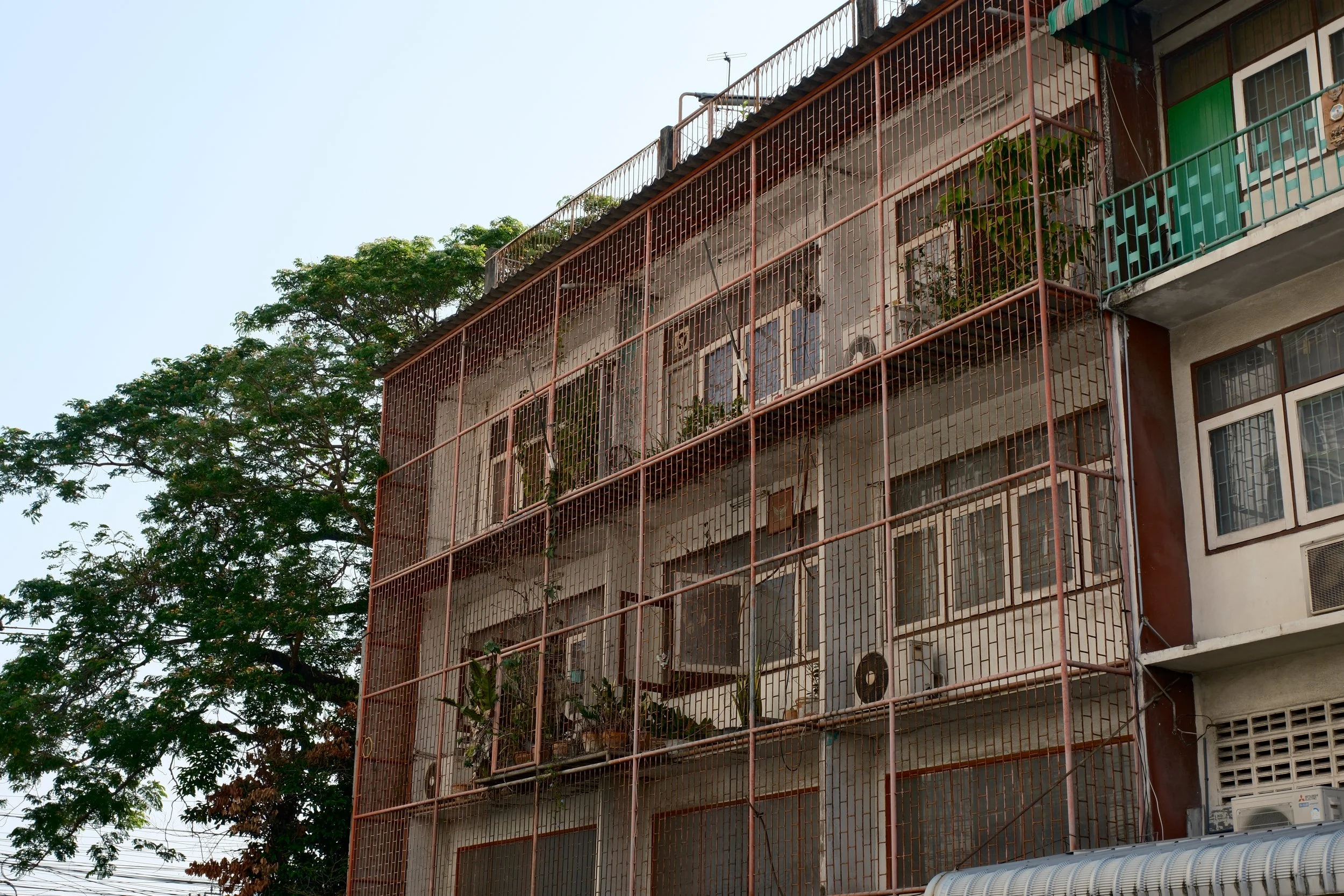 An apartment building with a metal security cage on the balconies, some of which have plants and air conditioning units, and a large tree beside it.
