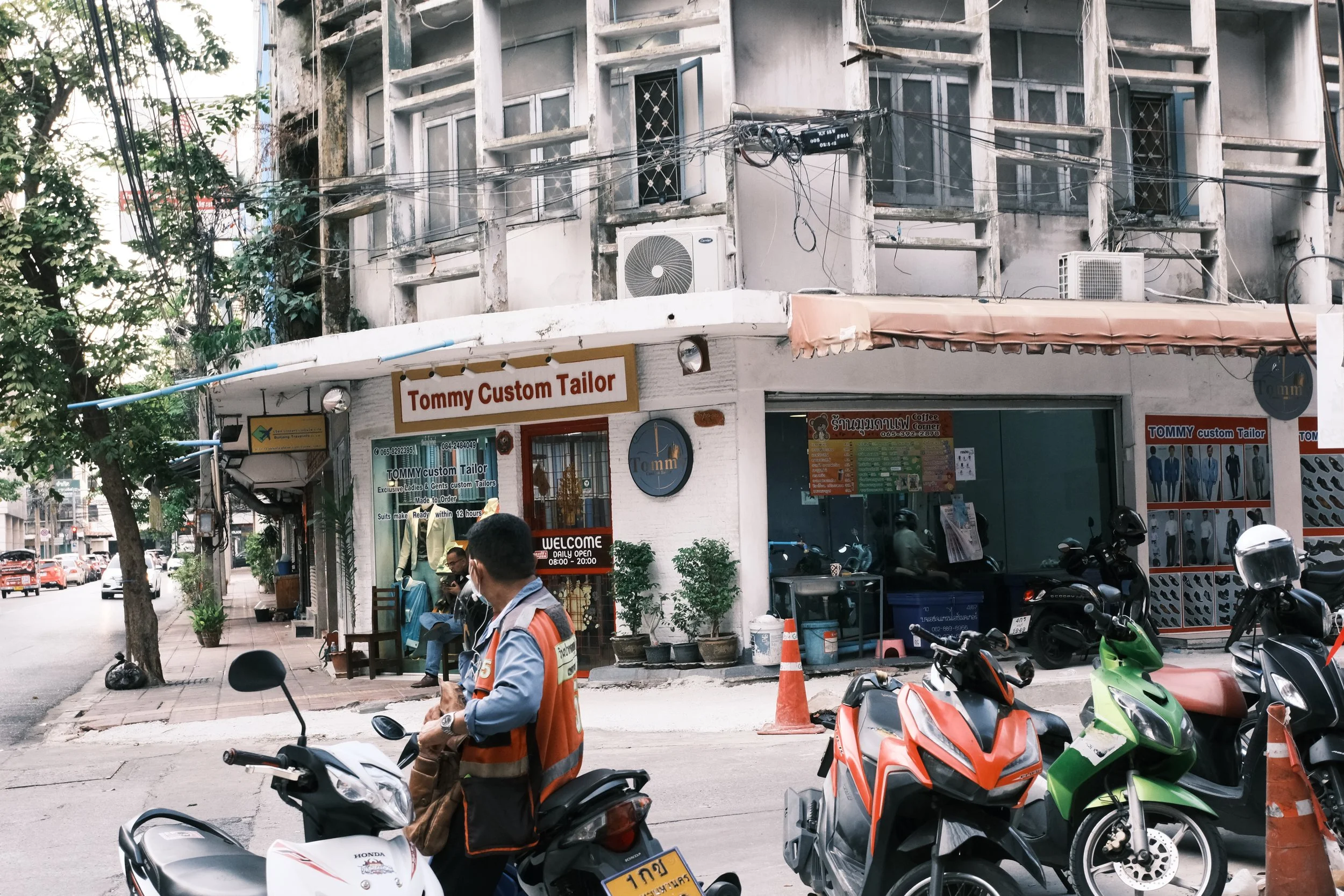 Street scene with a motorcycle rider in an orange vest, parked motorcycles, and an unfinished building with air conditioning units above a tailor shop named Tommy Custom Tailor.