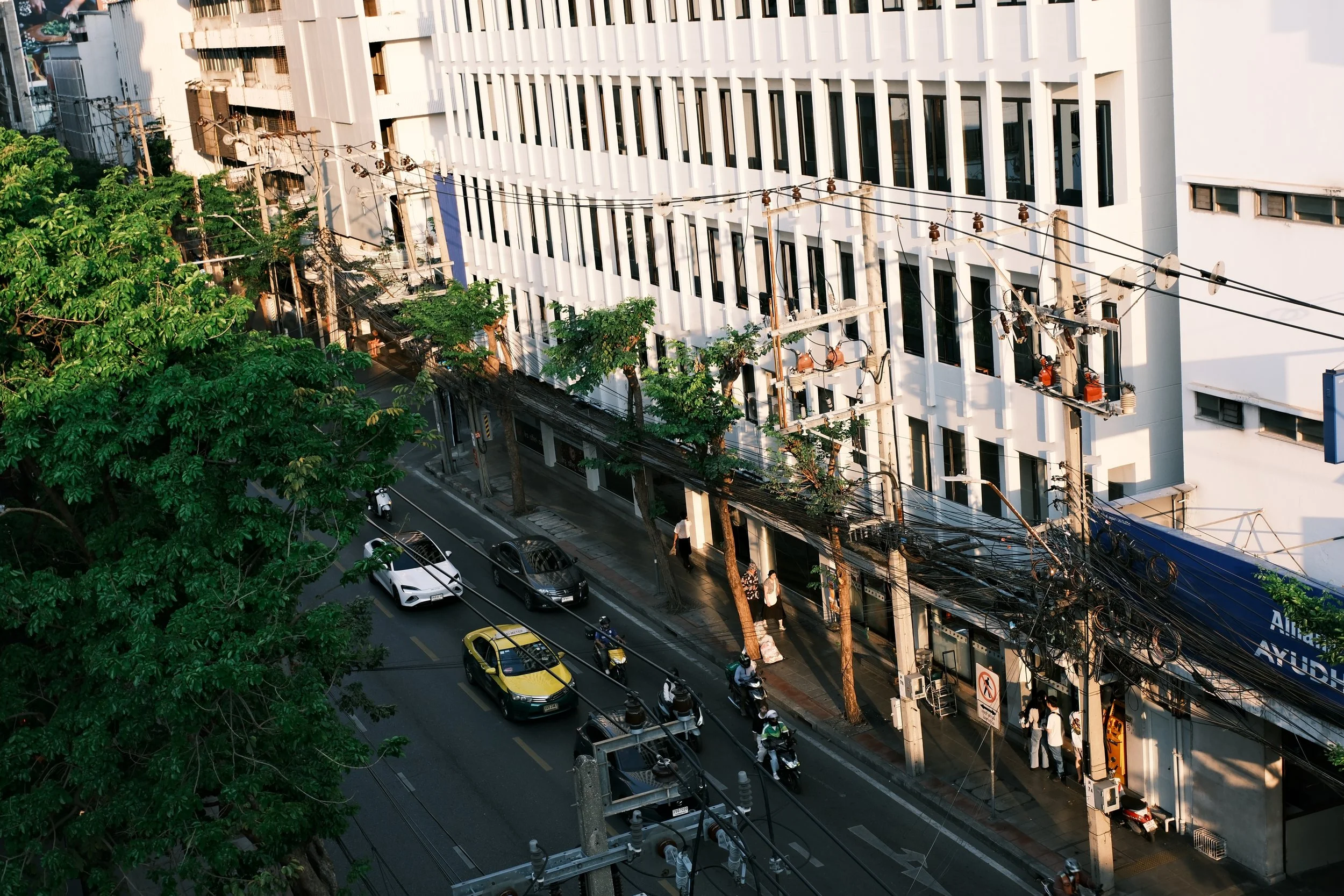 An urban street scene with a white modern building, trees, electrical wires, and vehicles including cars and motorcycles, with pedestrians on the sidewalk.