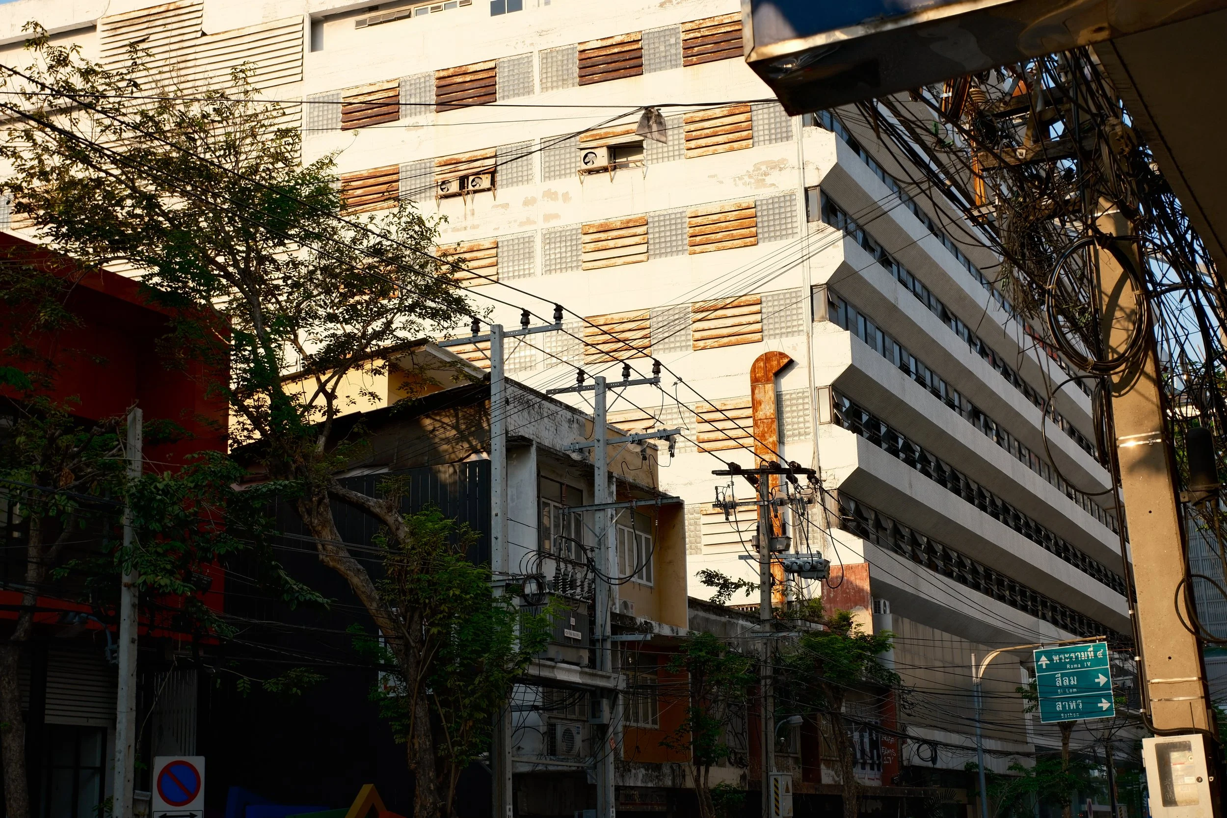 Urban street scene with a large building featuring textured wall panels, trees, power lines, and street signs in an Asian city.