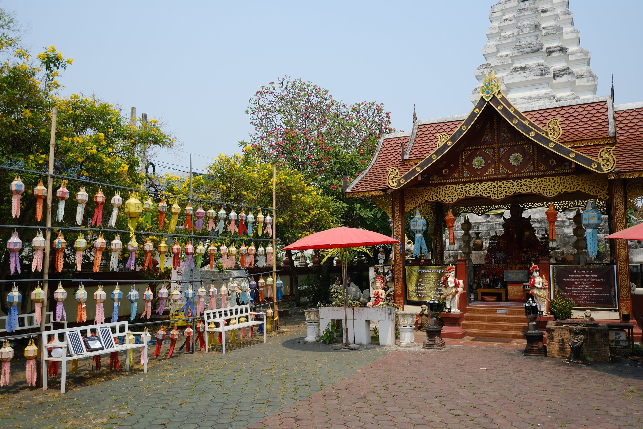 A traditional Thai temple with a decorated roof, statues, and signs, surrounded by colorful paper lanterns hanging on a fence, with white benches, pink umbrellas, and trees in the background.