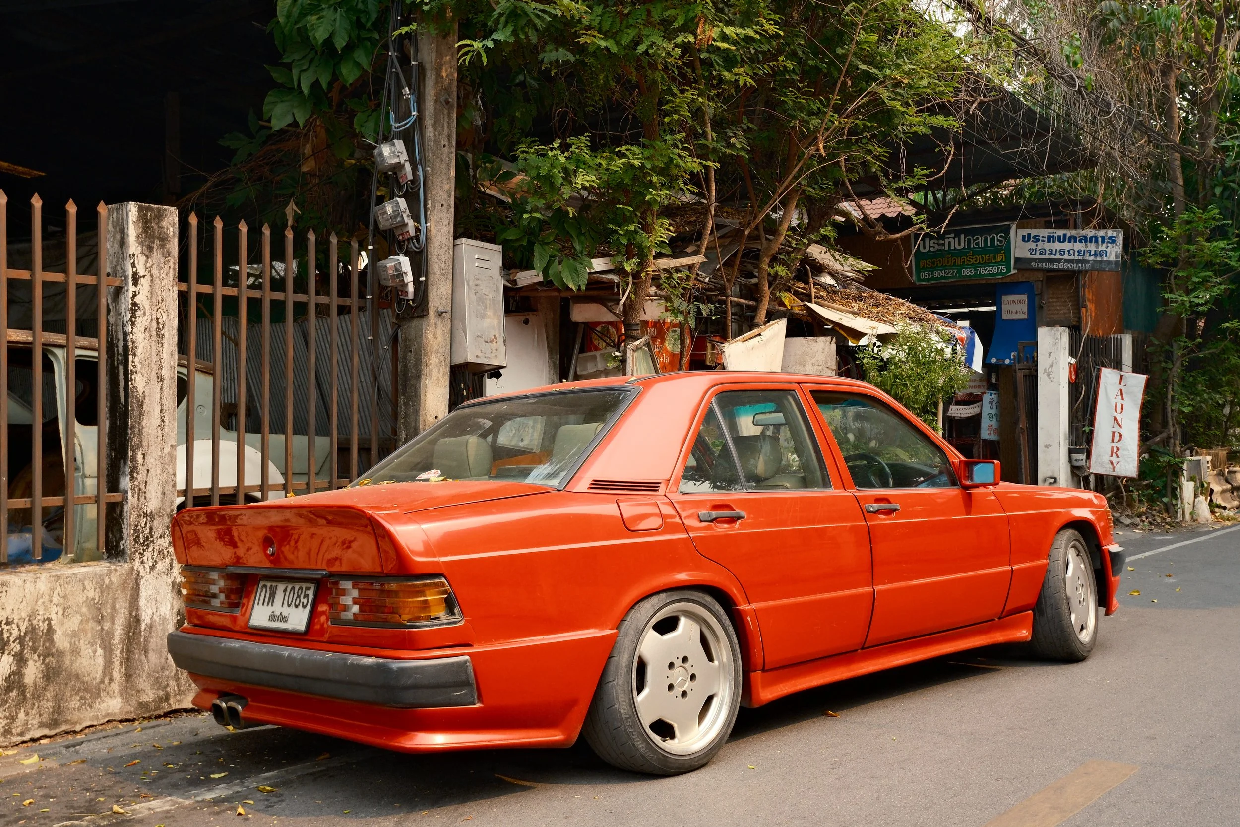 A vintage orange sedan parked beside a concrete curb on a street in front of a rustic building with various signs, trees, and foliage in the background.