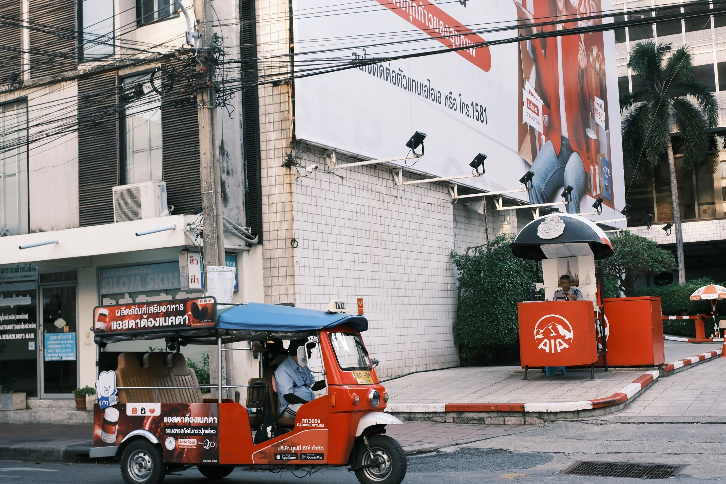 Street scene with a red tuk-tuk, a guard booth with AIA logo, large billboard, and a man sitting inside the booth