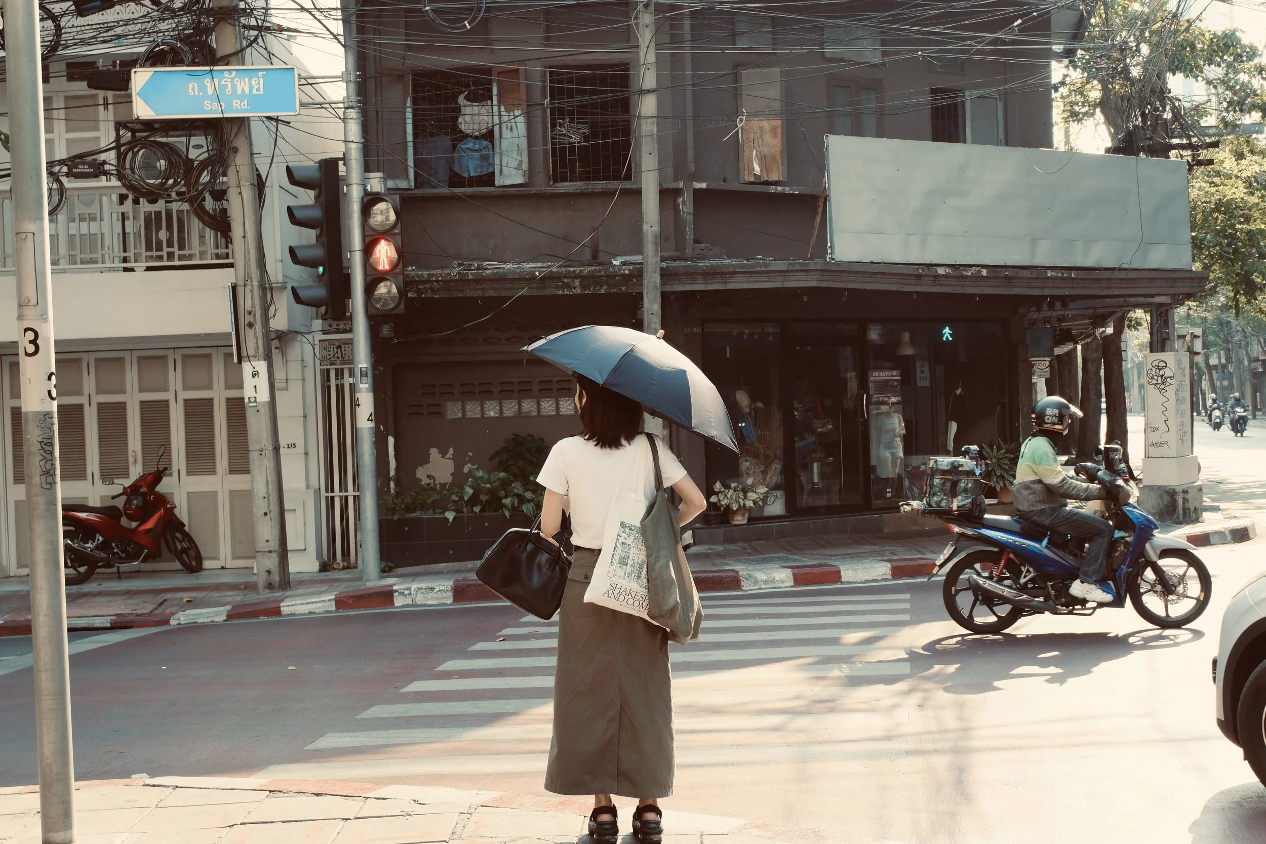A woman standing at a crosswalk holding an umbrella, with a motorcycle and an older building in the background and a traffic light showing red.