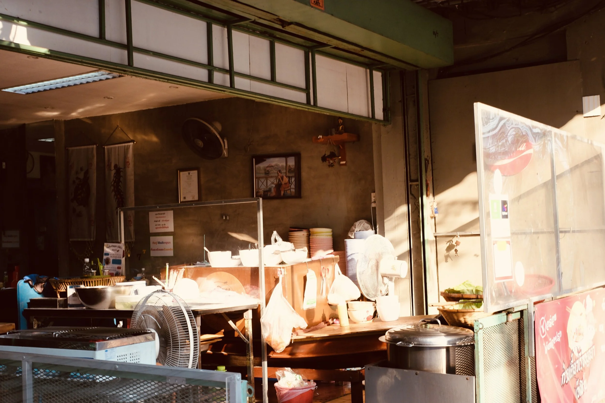 Sunlight illuminating the exterior of a small restaurant or food stall with various kitchen items, plates, and a fan visible inside.