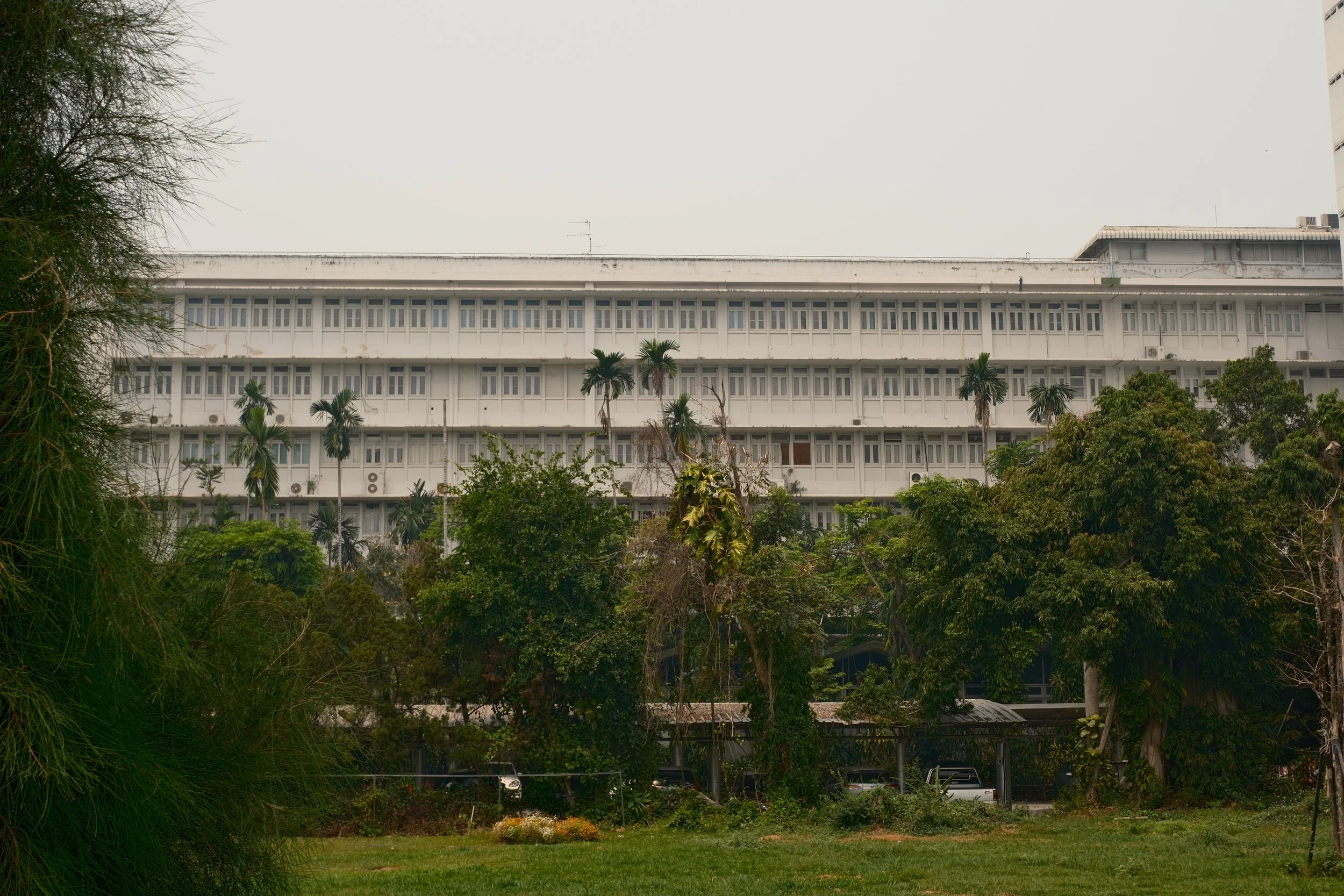 A large white building with multiple floors and many windows, surrounded by trees and greenery, under an overcast sky.