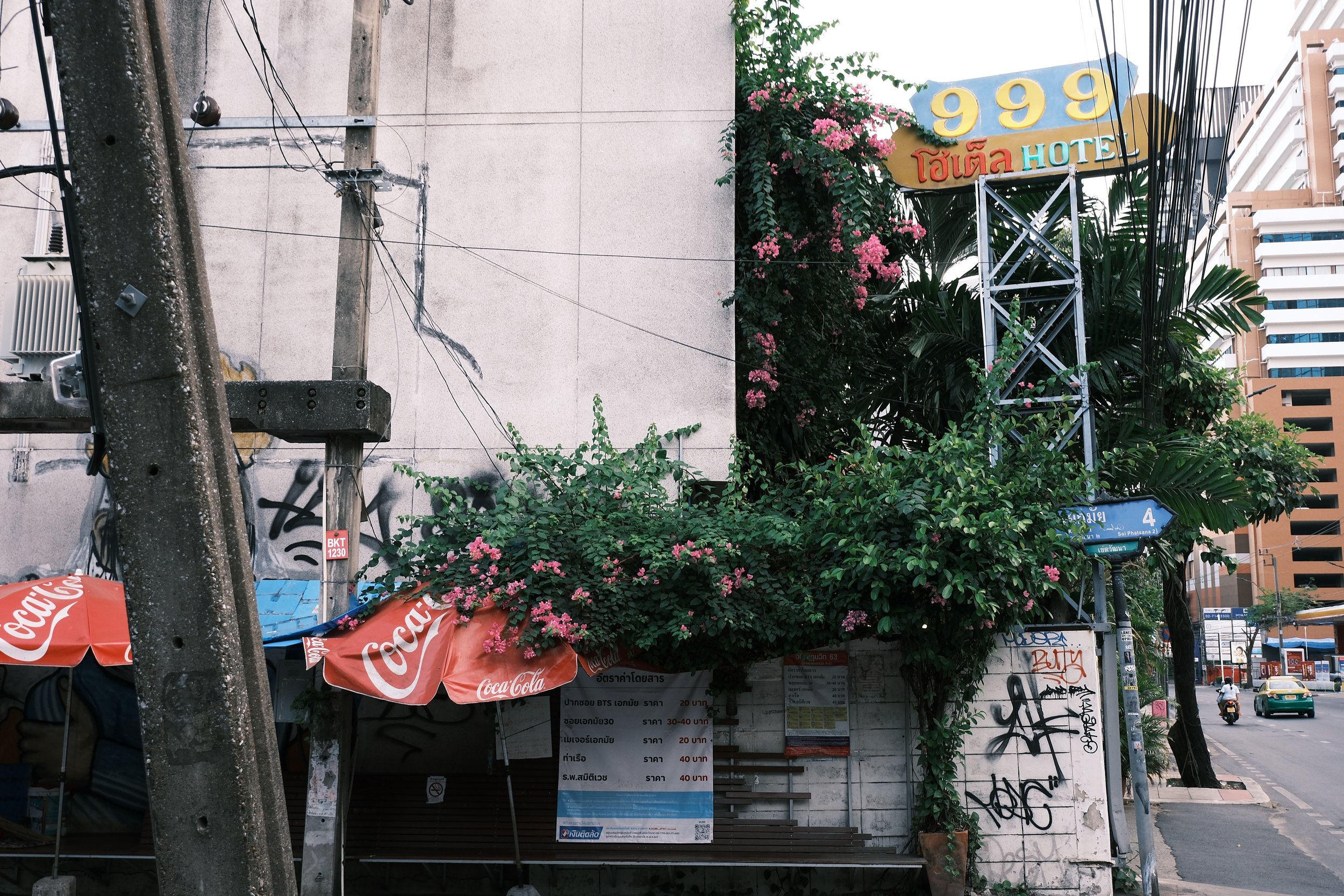 City street scene with a hotel sign, pink flowering plants, street signs, graffiti, and a roadside eatery with Coca-Cola umbrellas.