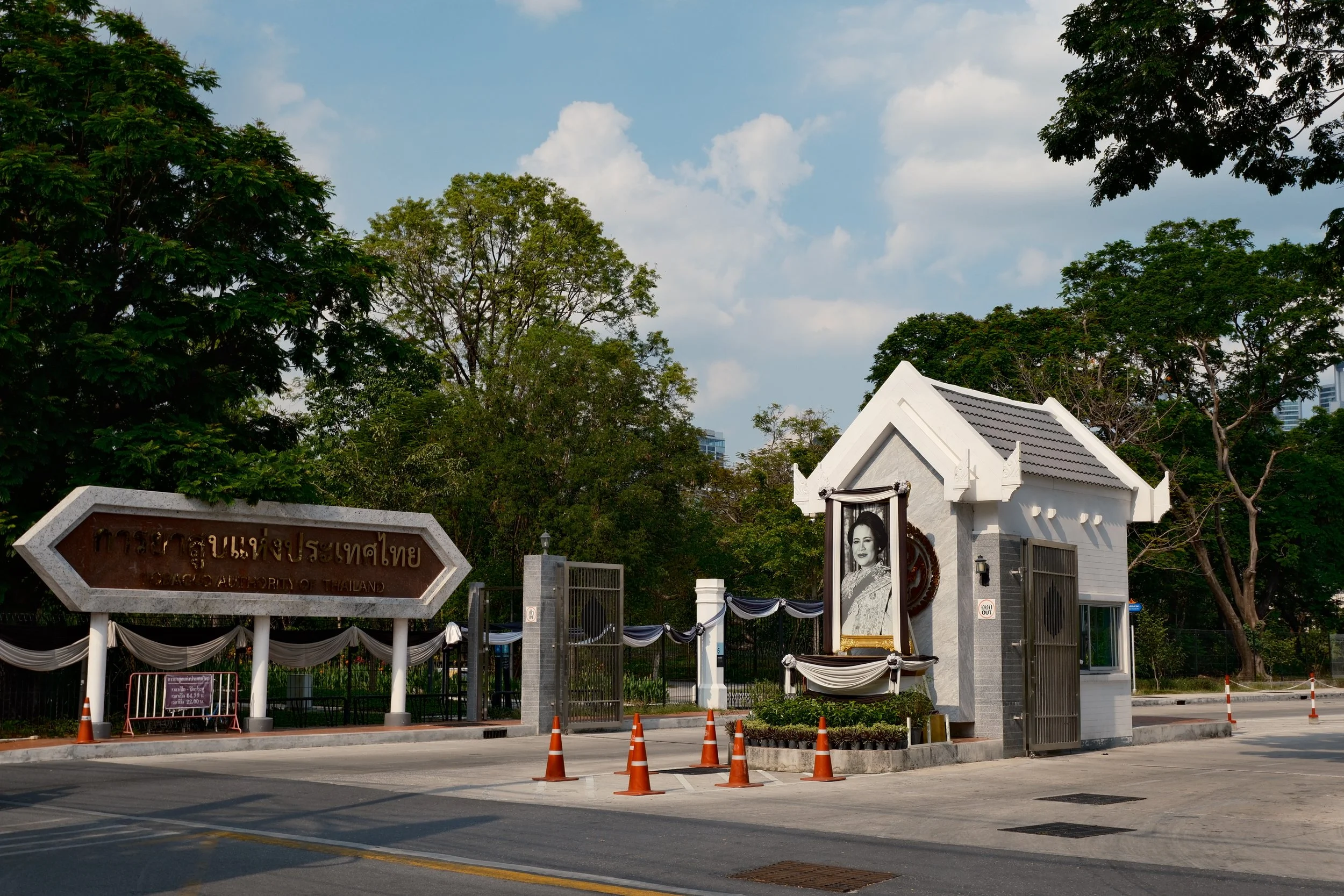 The image shows the entrance of the Office of the Prime Minister of Thailand, with a large portrait of a woman dressed in traditional Thai attire, and a sign with Thai and English text. The area is decorated with black and white fabric, and there are
