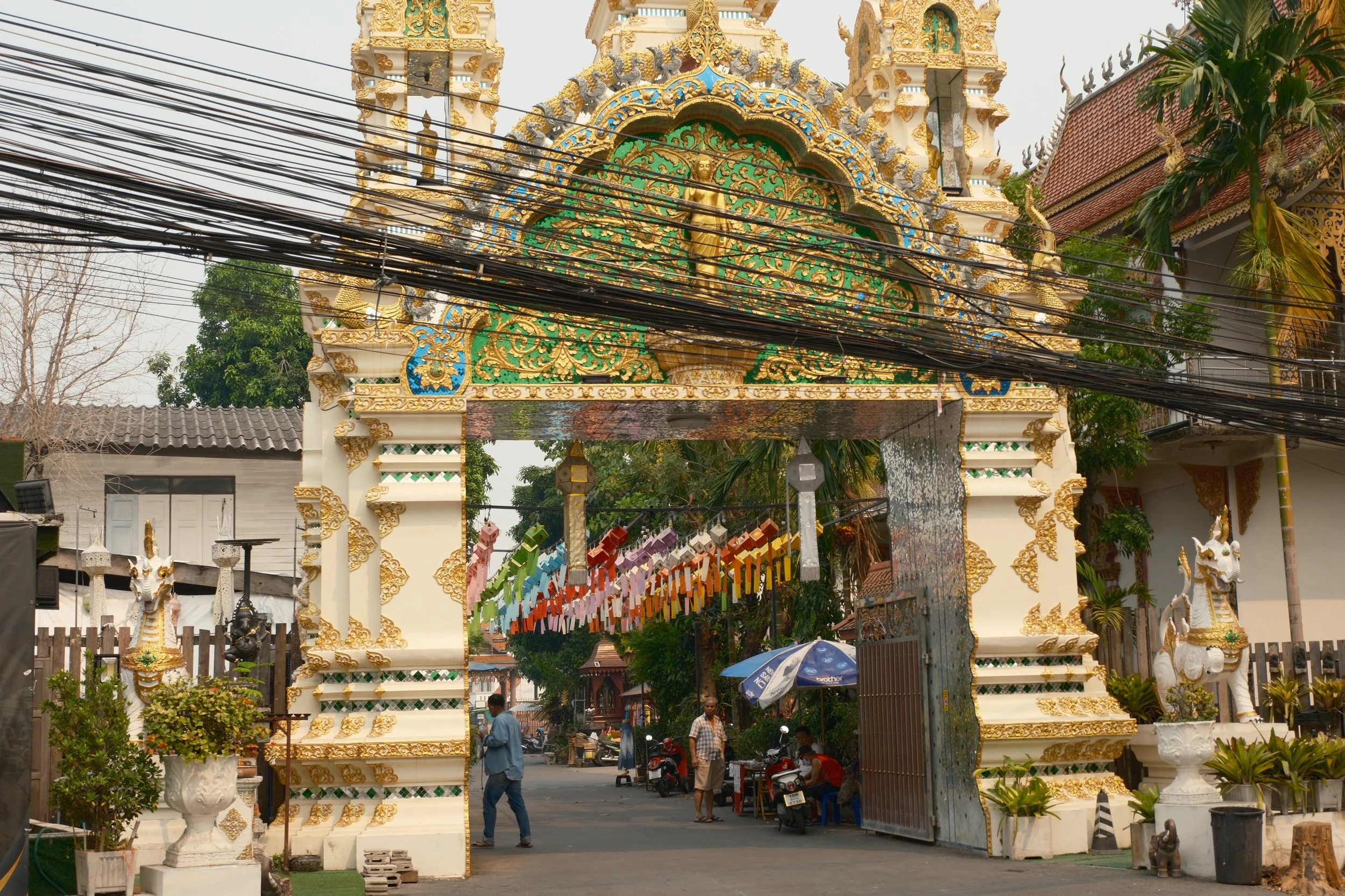 The entrance gate of a Buddhist temple, decorated with intricate gold and green designs, featuring statues of mythical creatures, and colorful paper lanterns hanging across the street. People walking and sitting nearby.