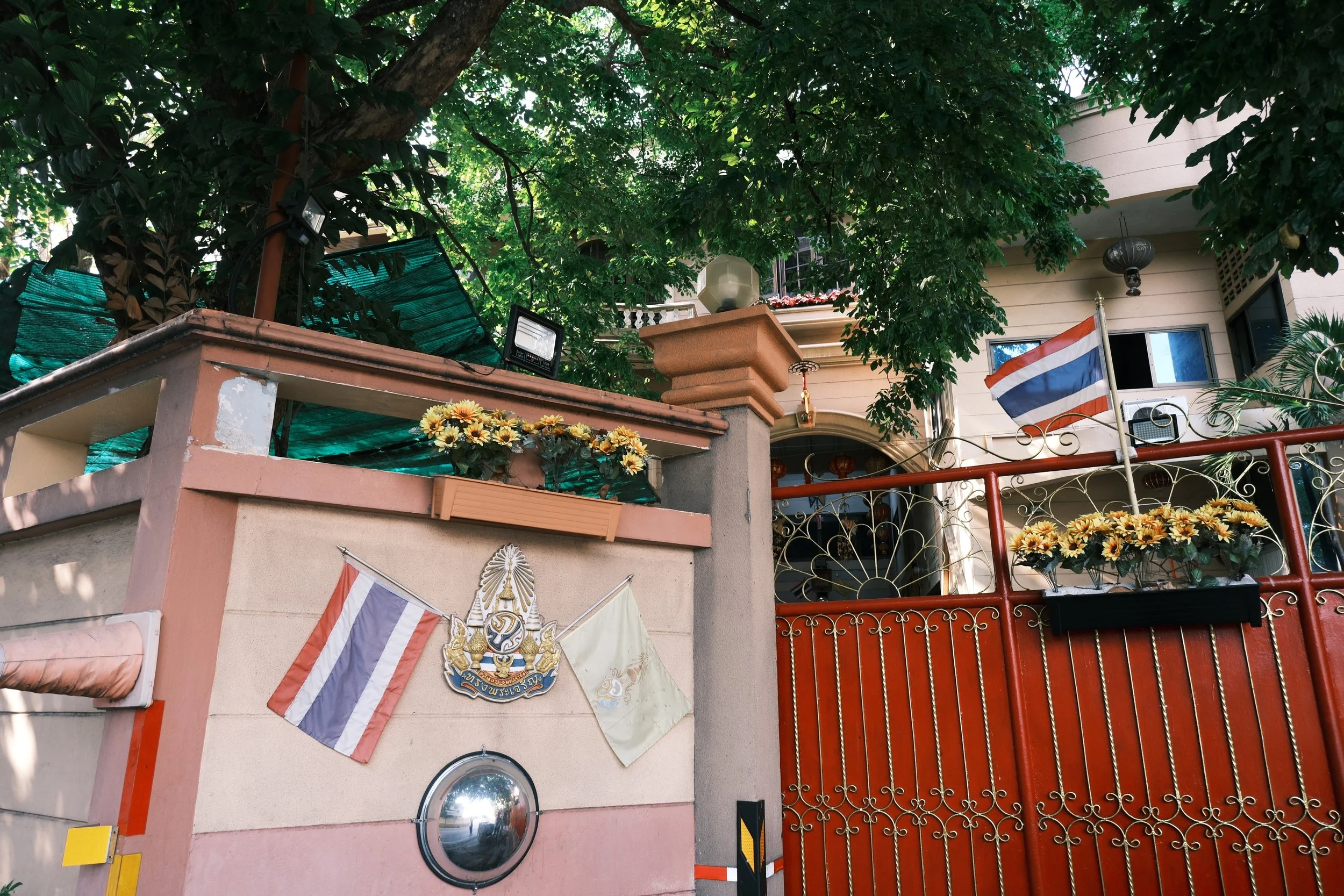 A house with a red metal gate and Thai flags, decorated with sunflowers and a crest, surrounded by greenery and trees.