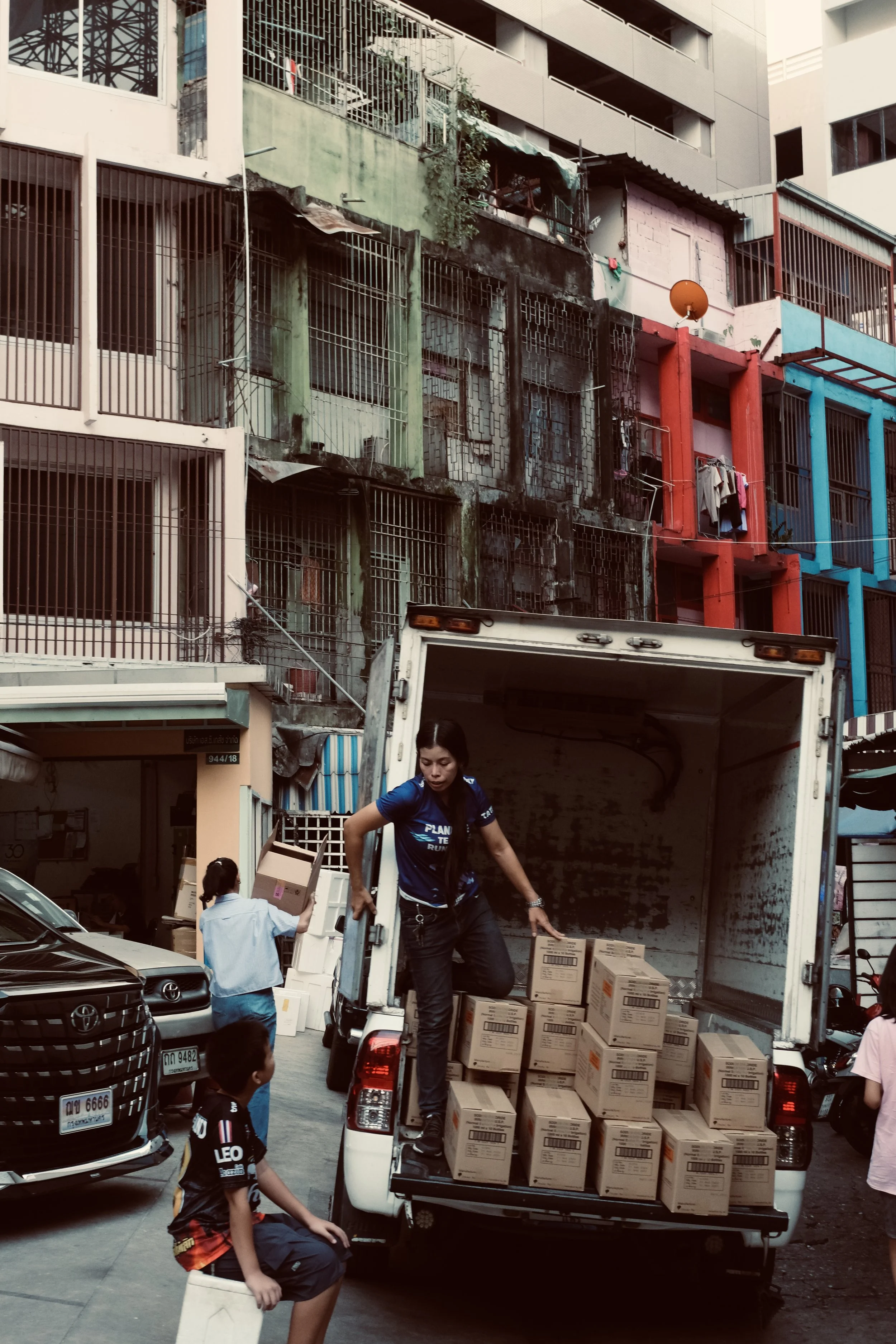A woman is unloading boxes from a truck onto a street surrounded by parked cars, with children nearby and colorful buildings with metal bars and balconies in the background.