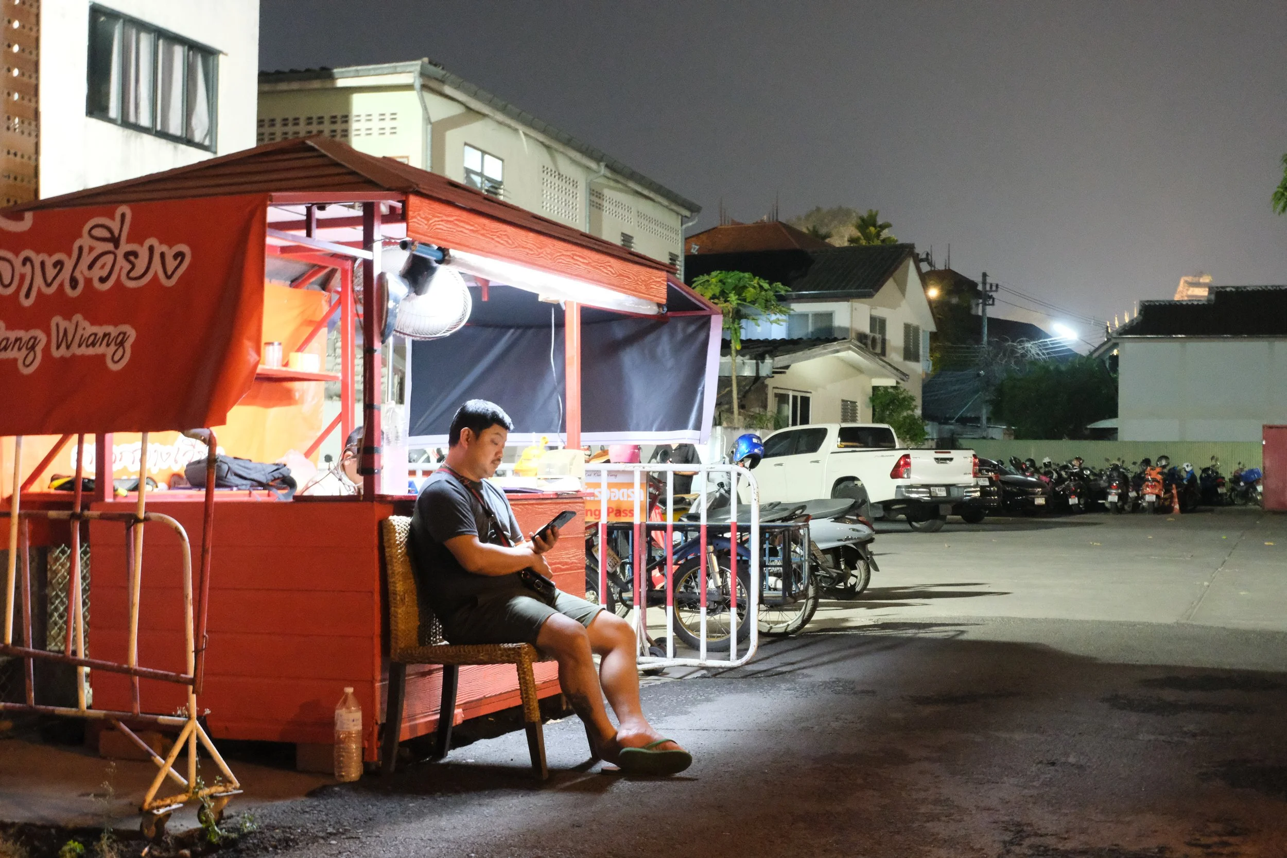 A man sitting outside a red food cart at night, looking at his phone, with buildings and parked cars in the background.