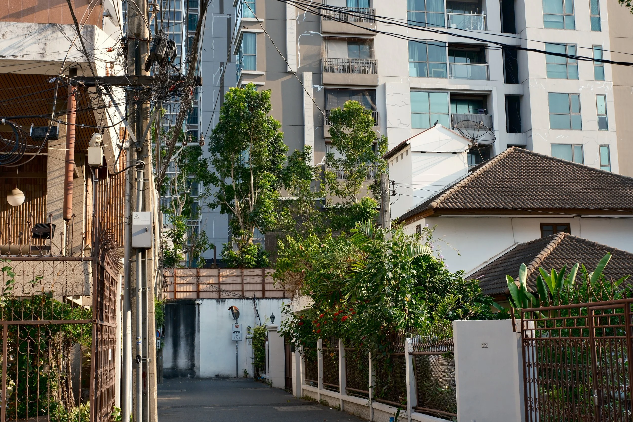 Urban scene with electrical poles and wires, modern apartment building, and a traditional house with a tiled roof, greenery, and fences.
