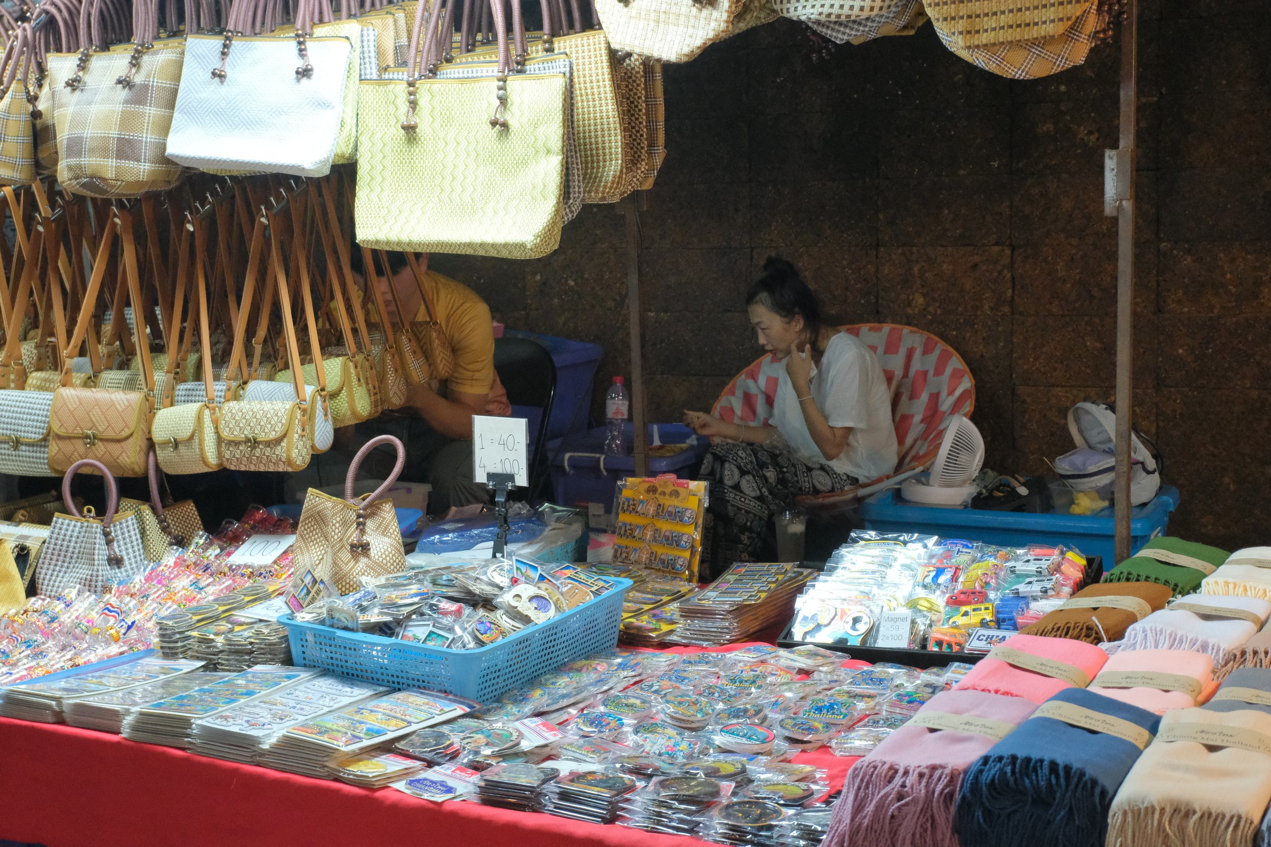 A marketplace stall with various bags, keychains, and souvenirs displayed on tables and hanging from above. Two women are behind the stall, one sitting and looking at her phone.