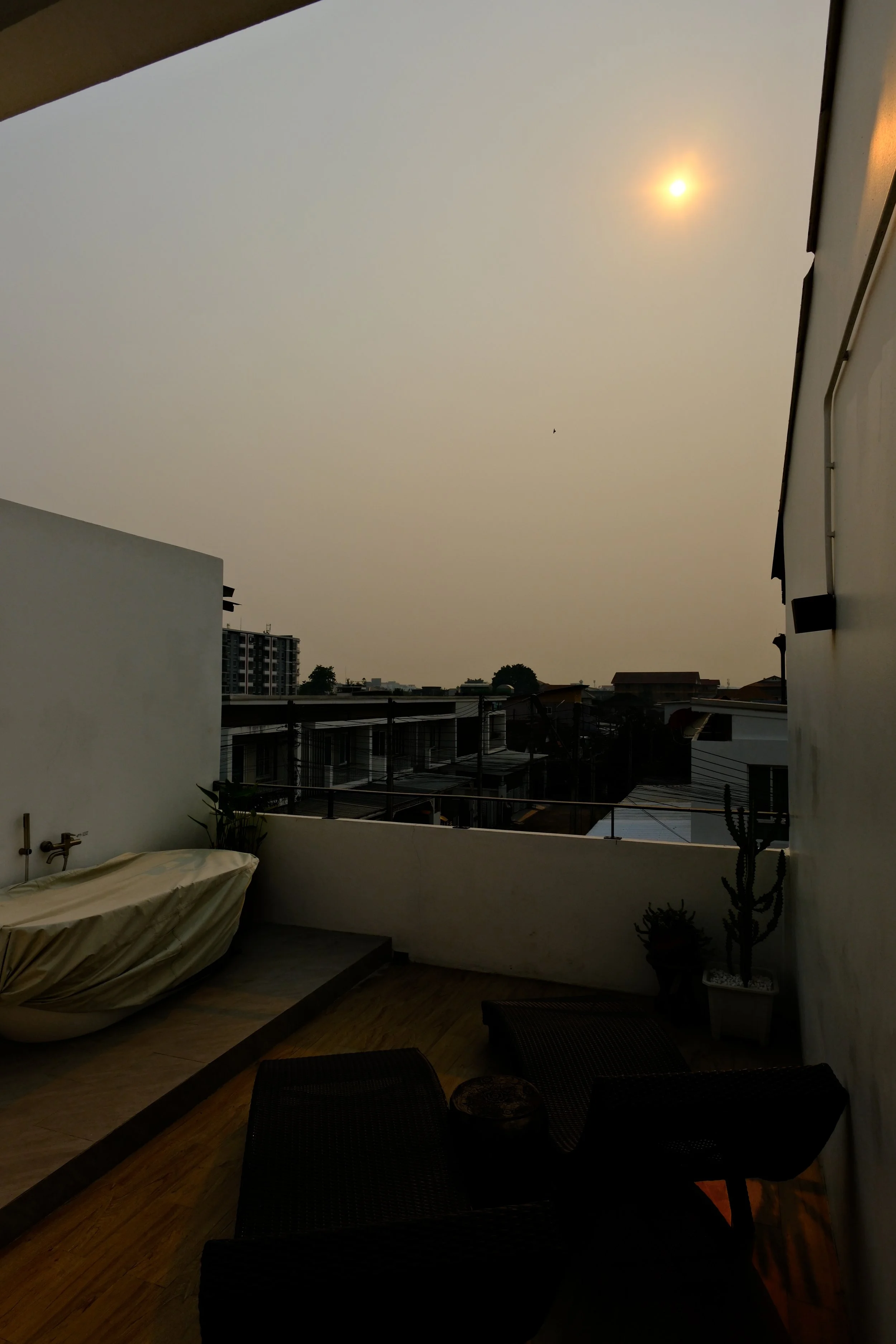 A rooftop patio with outdoor furniture, potted plants, and a covered bathtub, overlooking a cityscape under a hazy sky with the sun partially obscured.