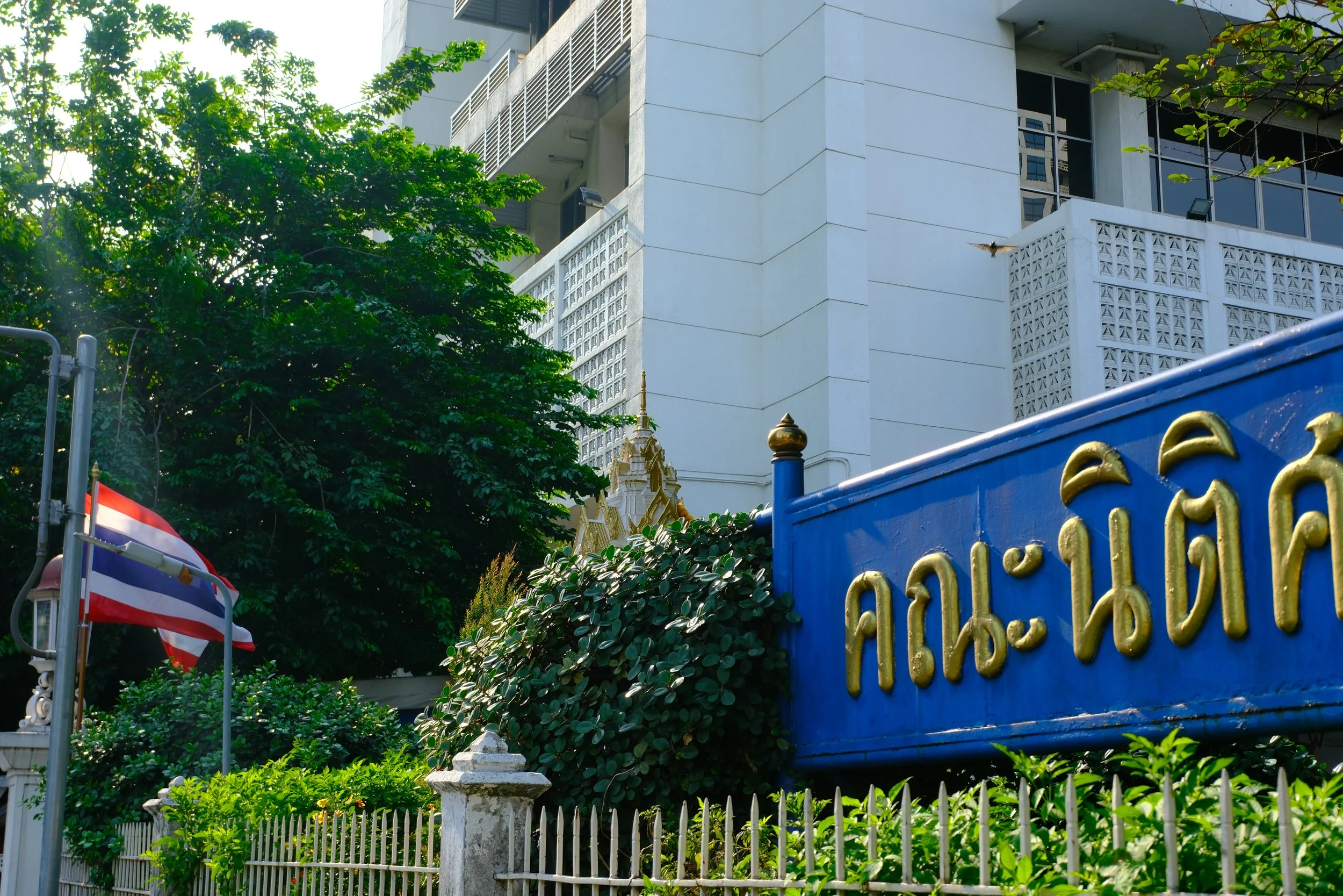 View of a modern white building with greenery in the foreground, including a flag on a pole and a blue sign with gold lettering in a foreign language.
