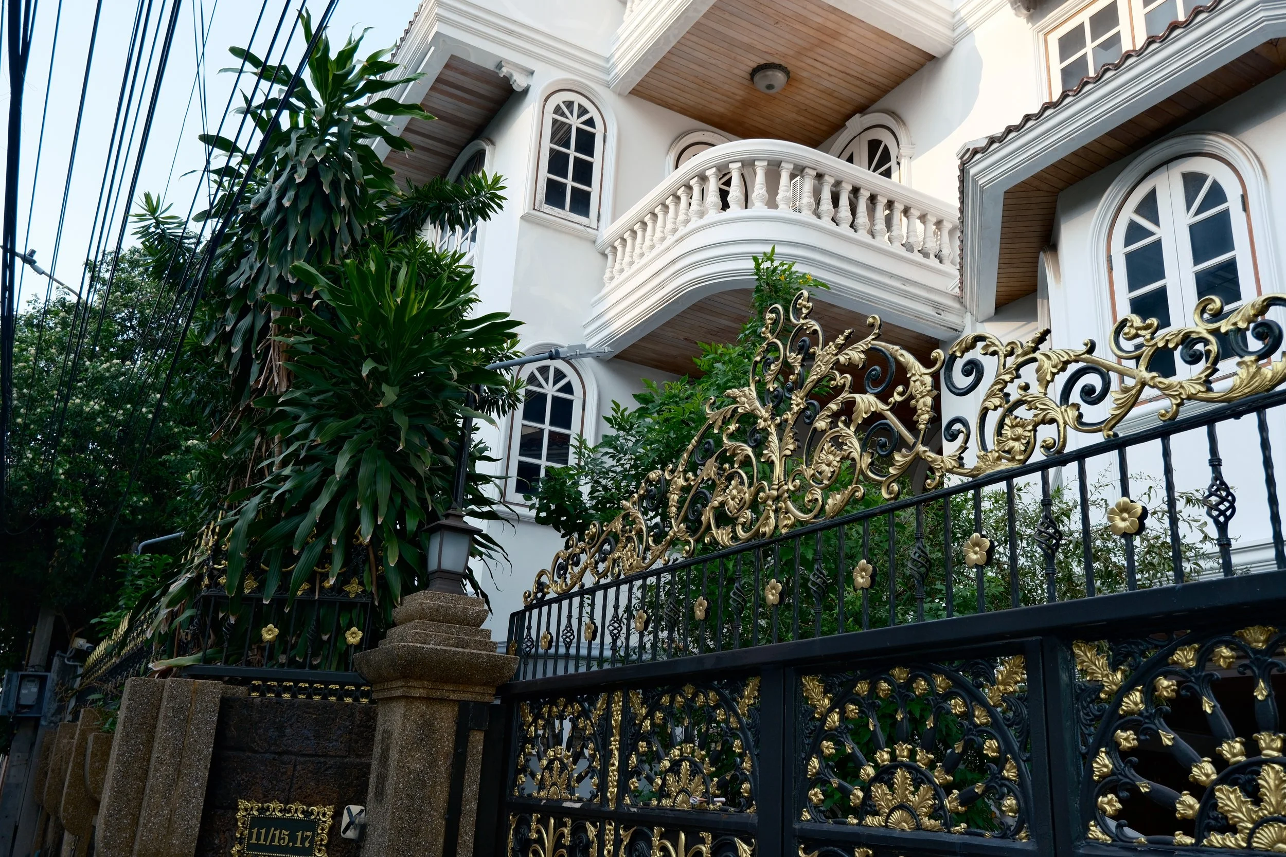 A white, multi-story house with arched windows, a balcony, and a decorative black and gold wrought iron gate in front, surrounded by green plants and trees.
