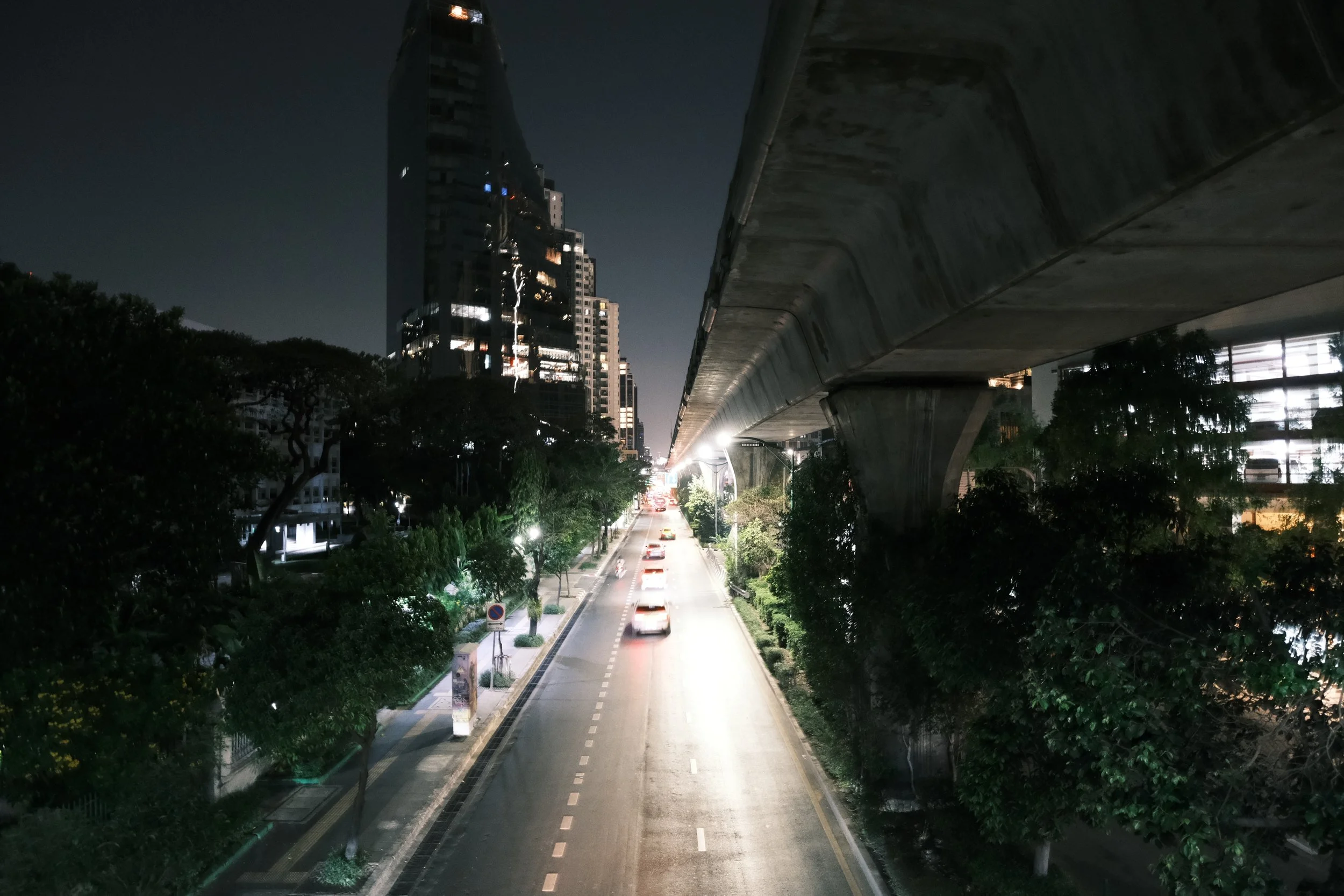 Night view of a city street with cars, trees, and high-rise buildings, featuring an elevated train track overhead.
