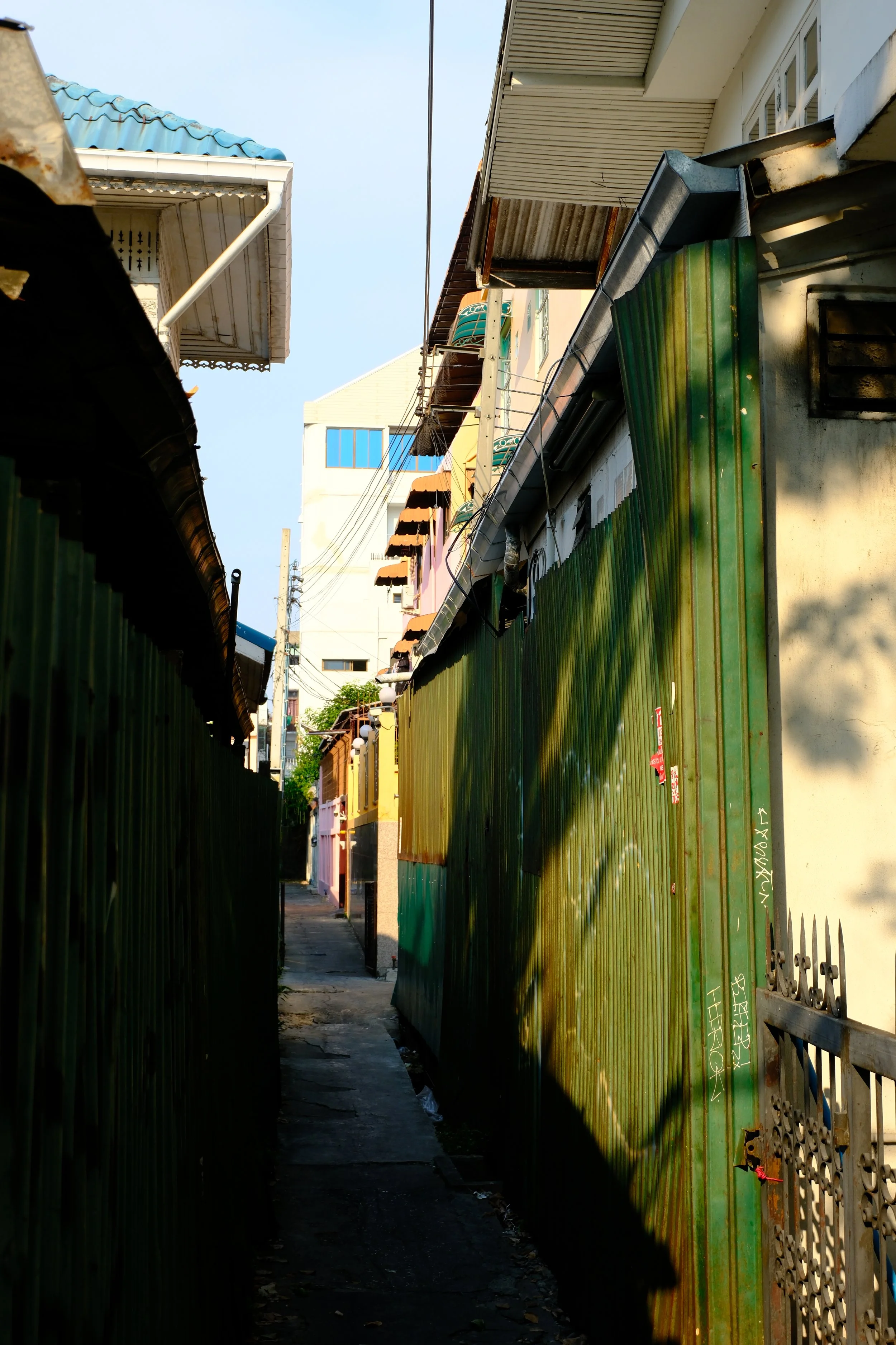 Narrow alleyway between brightly colored houses and green metal fences, with shadows of a tree cast on the fence on a sunny day.