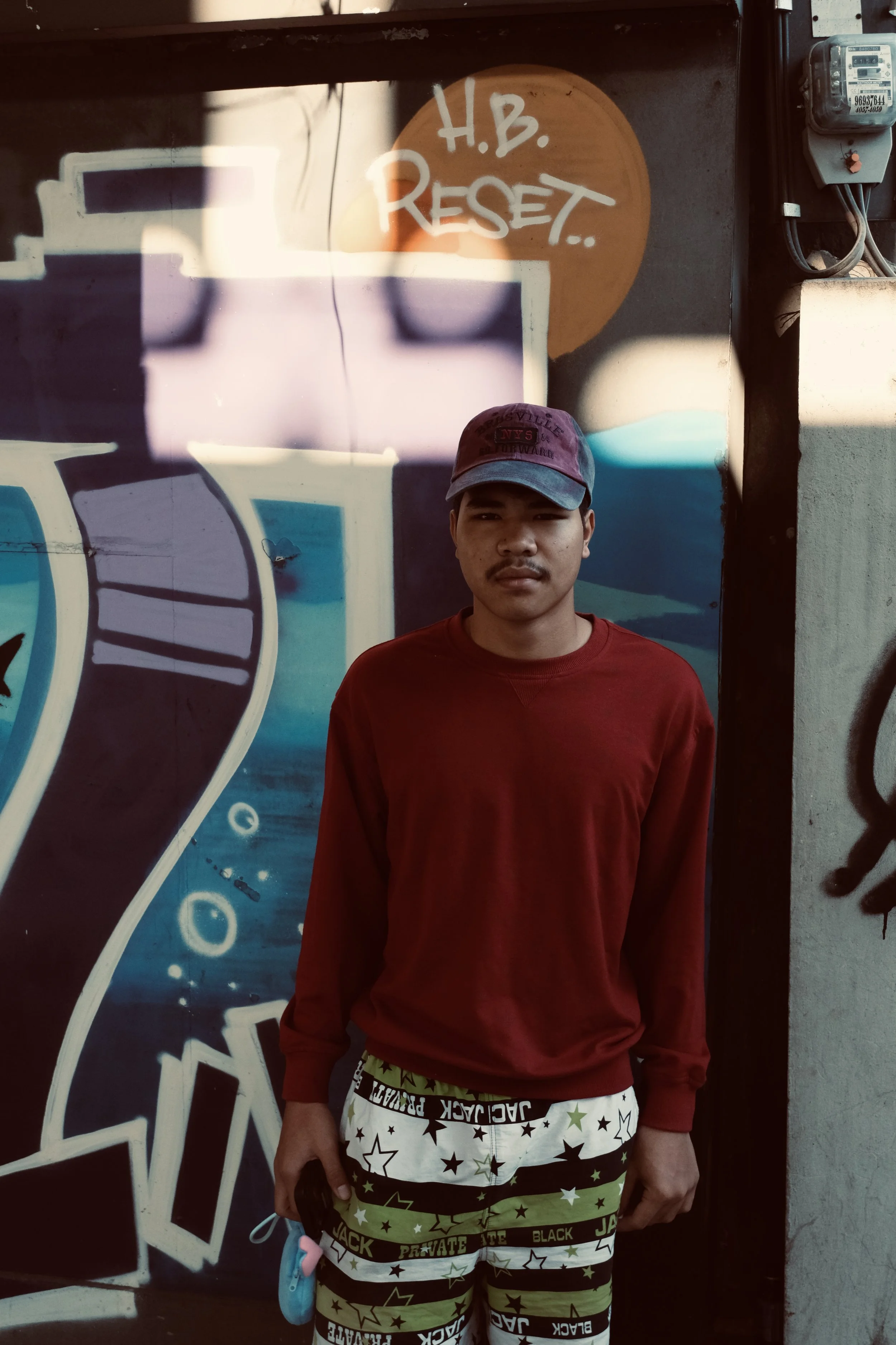 A young man with a mustache and goatee, wearing a burgundy sweatshirt, patterned shorts featuring stars and words, and a purple cap, standing in front of a graffiti-covered wall with the phrase 'H.B. REJECT' written in white. He is holding a small ob