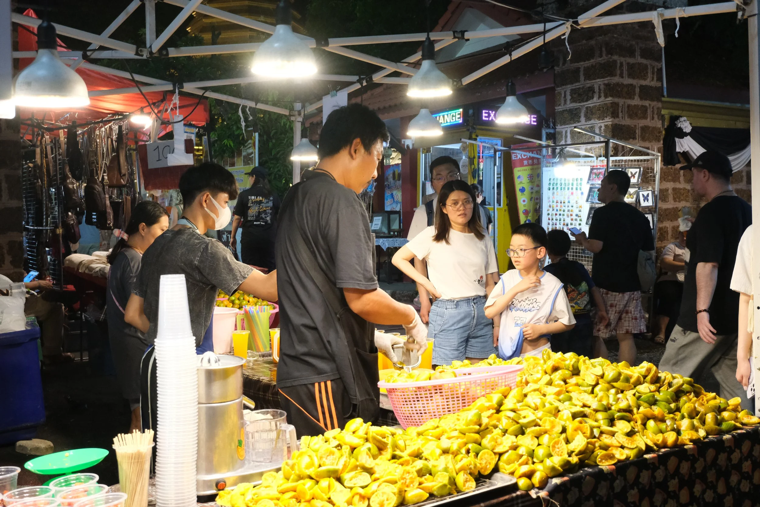 Night market scene with people around a lemon juice stall. Vendor preparing lemon drinks, pile of cut lemons on the table, some customers, and various stalls in the background illuminated with lights.