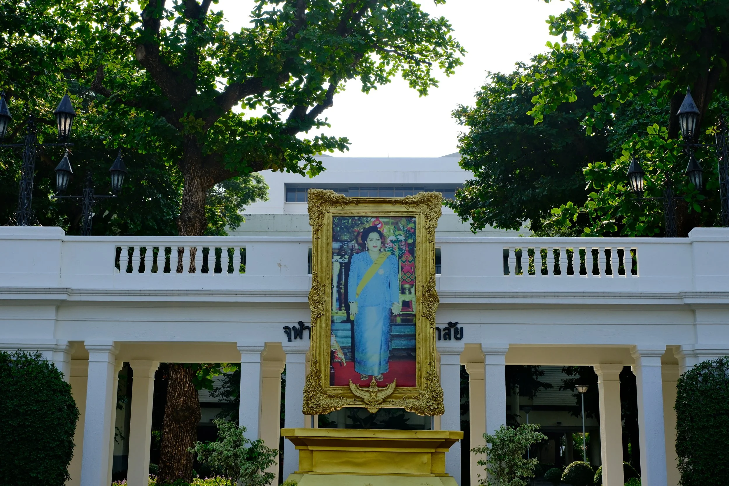 A large portrait of a woman in traditional Thai attire displayed in an ornate gold frame on a yellow pedestal, set against a white building with trees and black lamps in the background.