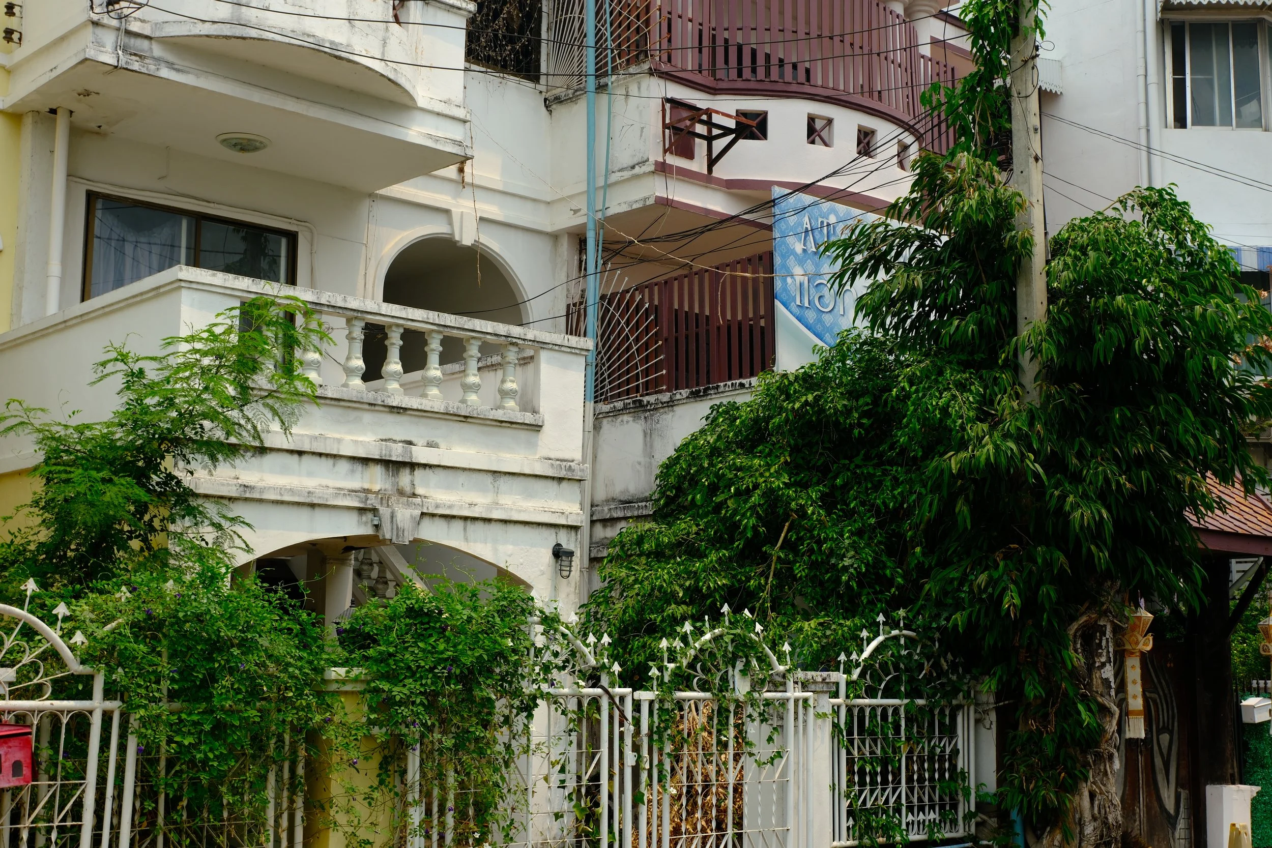 A white multi-story residential building with balconies, surrounded by lush green trees and a white metal fence.