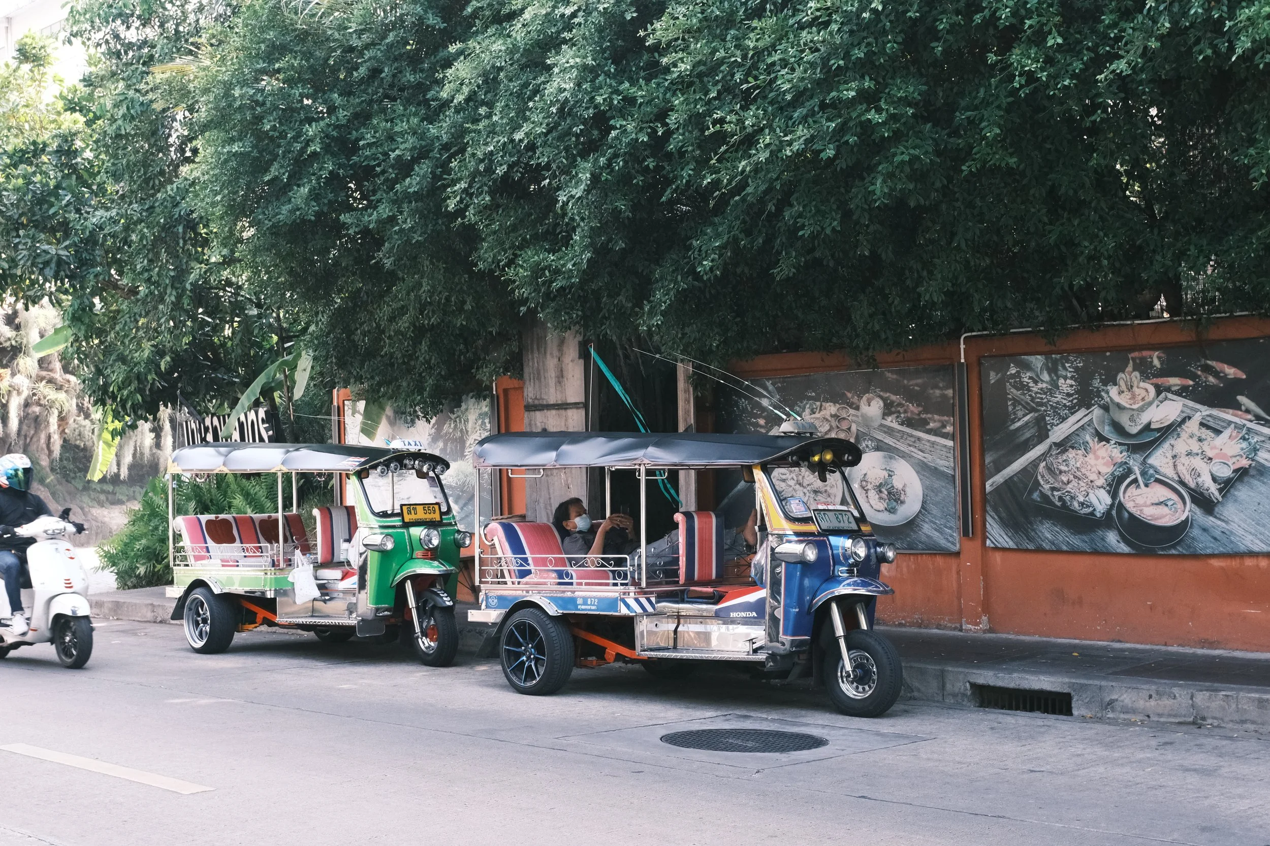 Two tuk-tuks with colorful seating parked on the side of a street under large green trees, with a person wearing a face mask sitting inside one, and a motorbike passing by on the road.