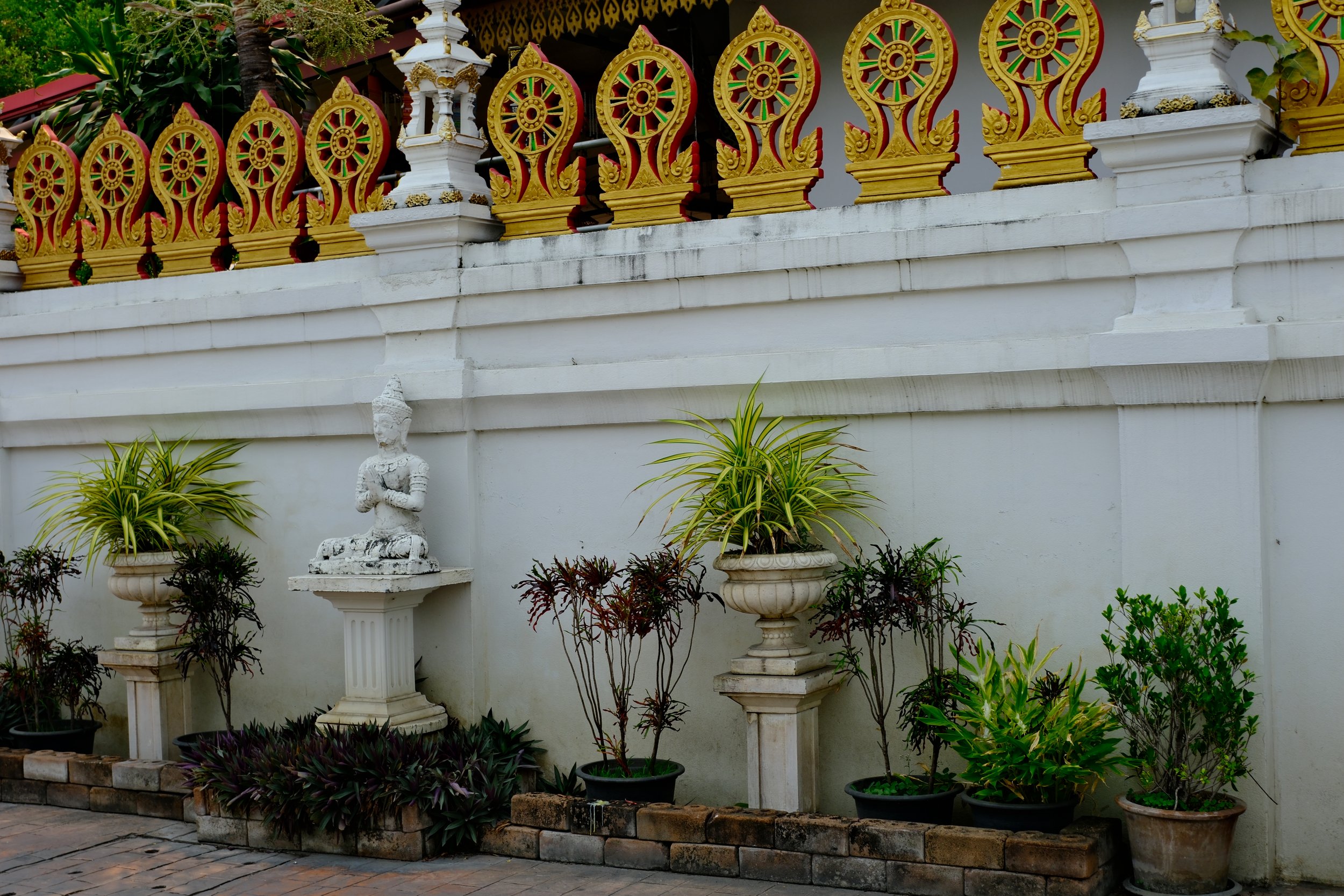 Decorative Thai-style temple railing with intricate gold and red patterns, white wall with a small white Buddha statue sitting on a pedestal, surrounded by potted plants and shrubs.