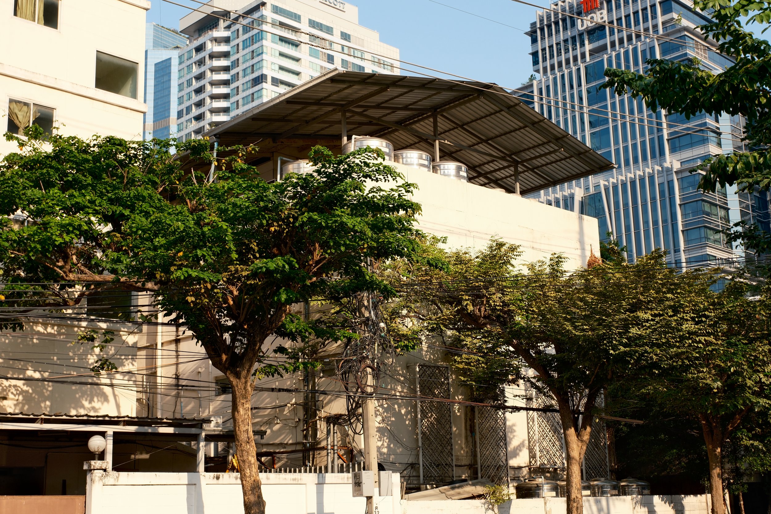 Cityscape with trees in foreground, older white building with metal roof and air conditioning units, modern glass skyscrapers in background.