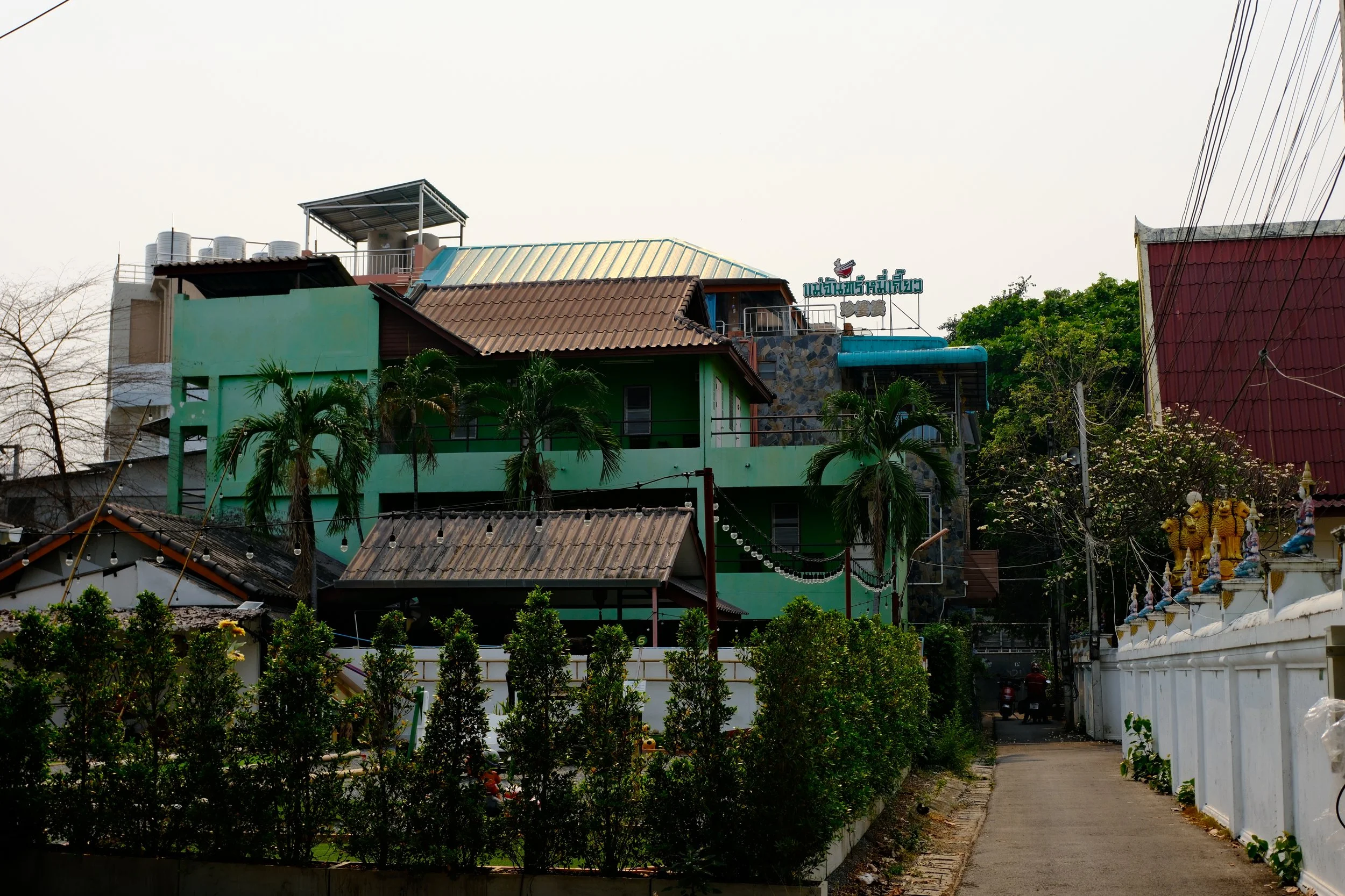 A colorful multi-story building with green exterior, palm trees, a narrow walkway, and decorative statues along the white wall on the right side.