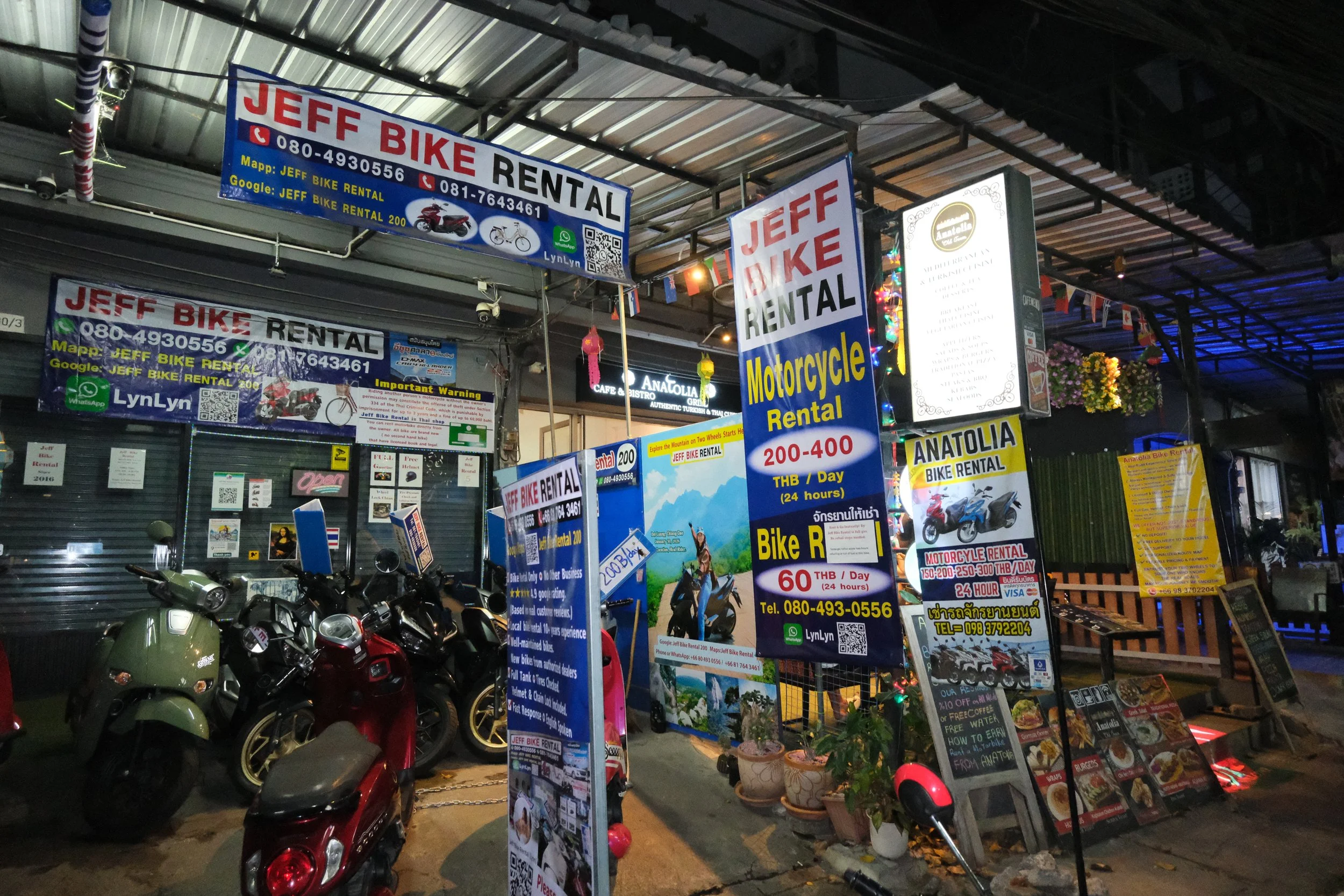 Street scene at night showing signs for Jeep Bike Rental and Anatolia Bike Rental with motorcycles parked in front, colorful lights, and restaurant menus displayed on the sidewalk.