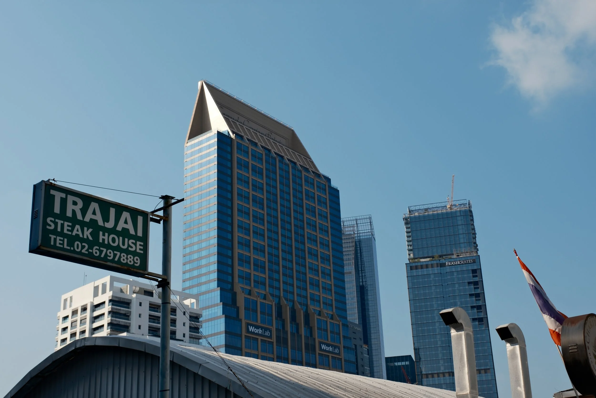 City skyline with modern skyscrapers against a blue sky, featuring a sign for Trajaj Steak House and a partially visible flag on the right.