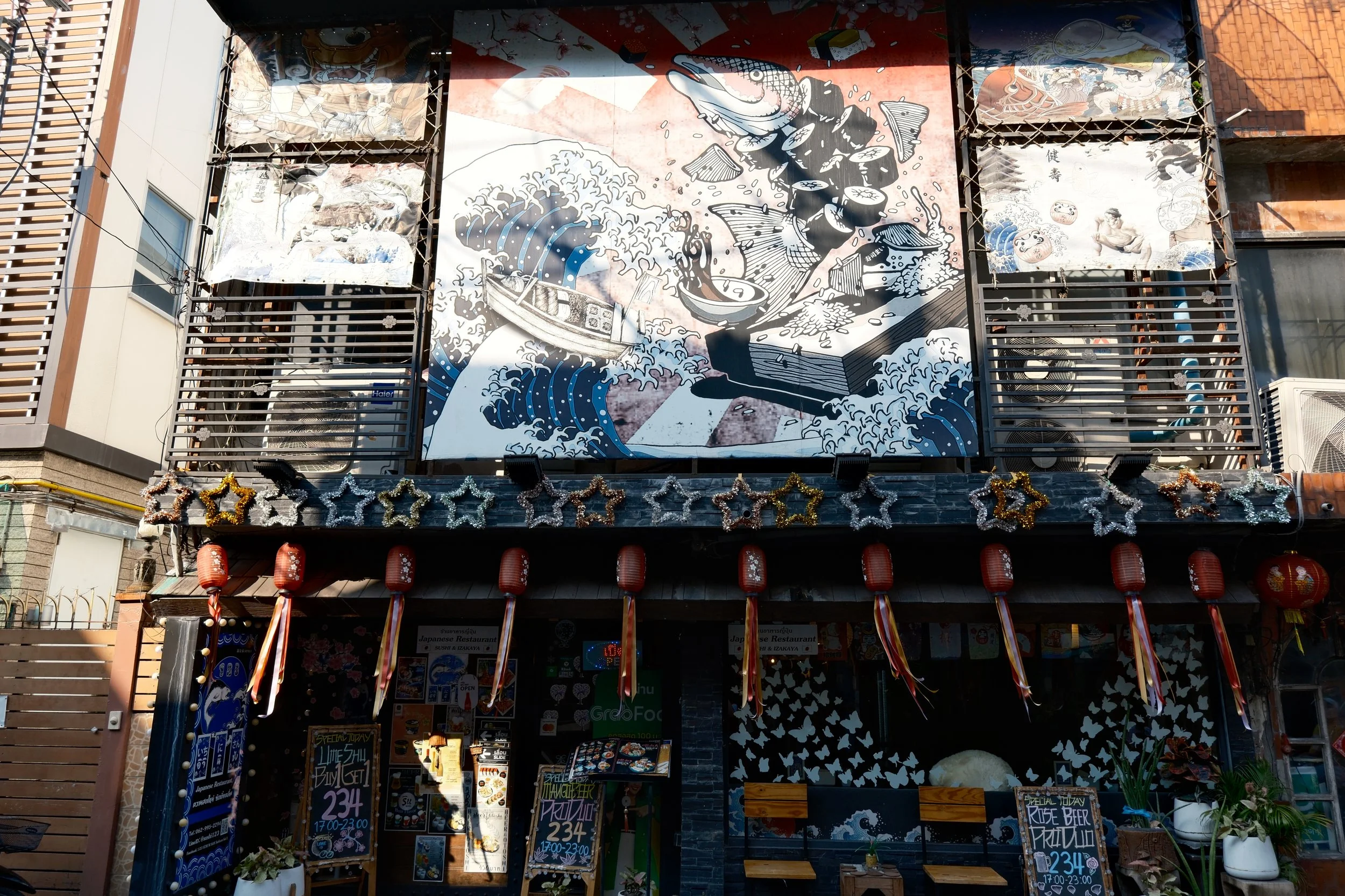 A building with a large mural depicting a scene from Japanese culture, with waves, boats, and fish, above the entrance decorated with lanterns and star-shaped ornaments.