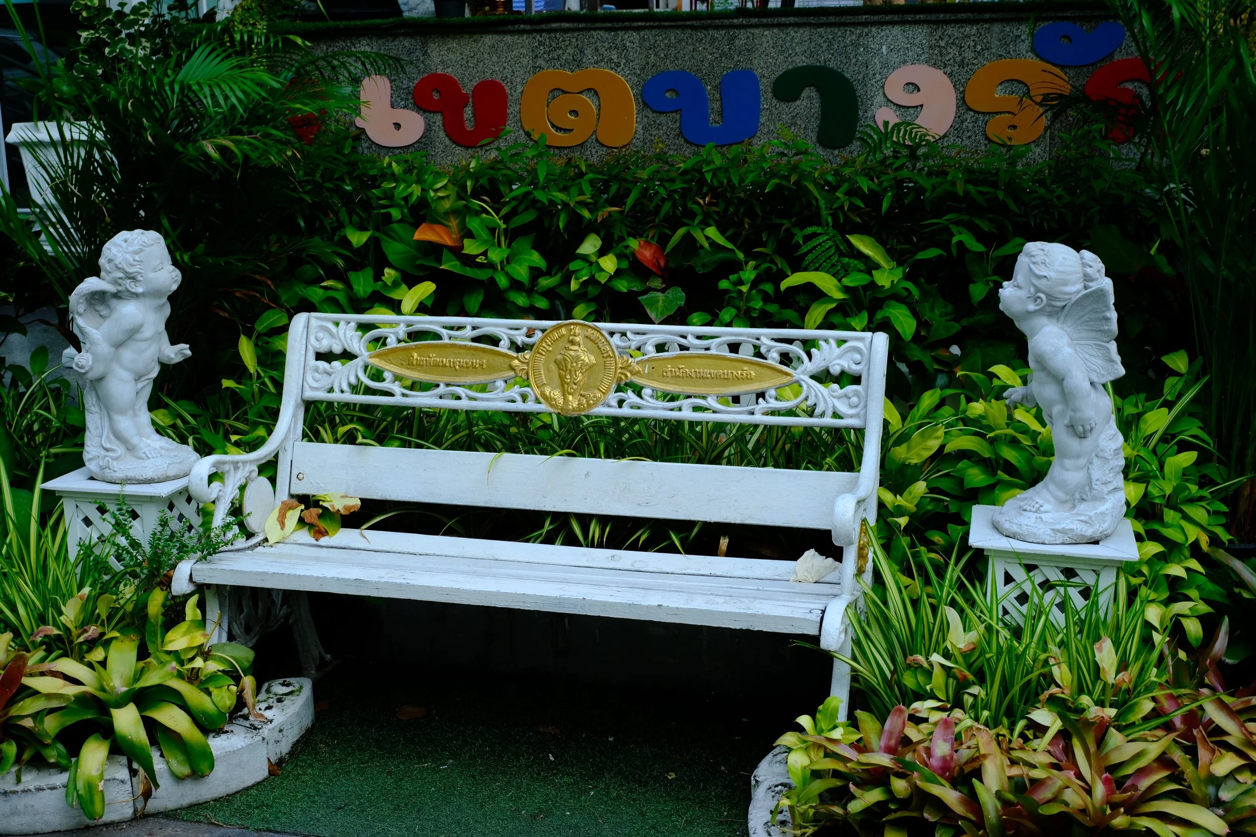 A white ornate metal bench with gold accents, flanked by two white statues of children in a garden with lush green plants. A colorful sign with Thai characters is in the background.