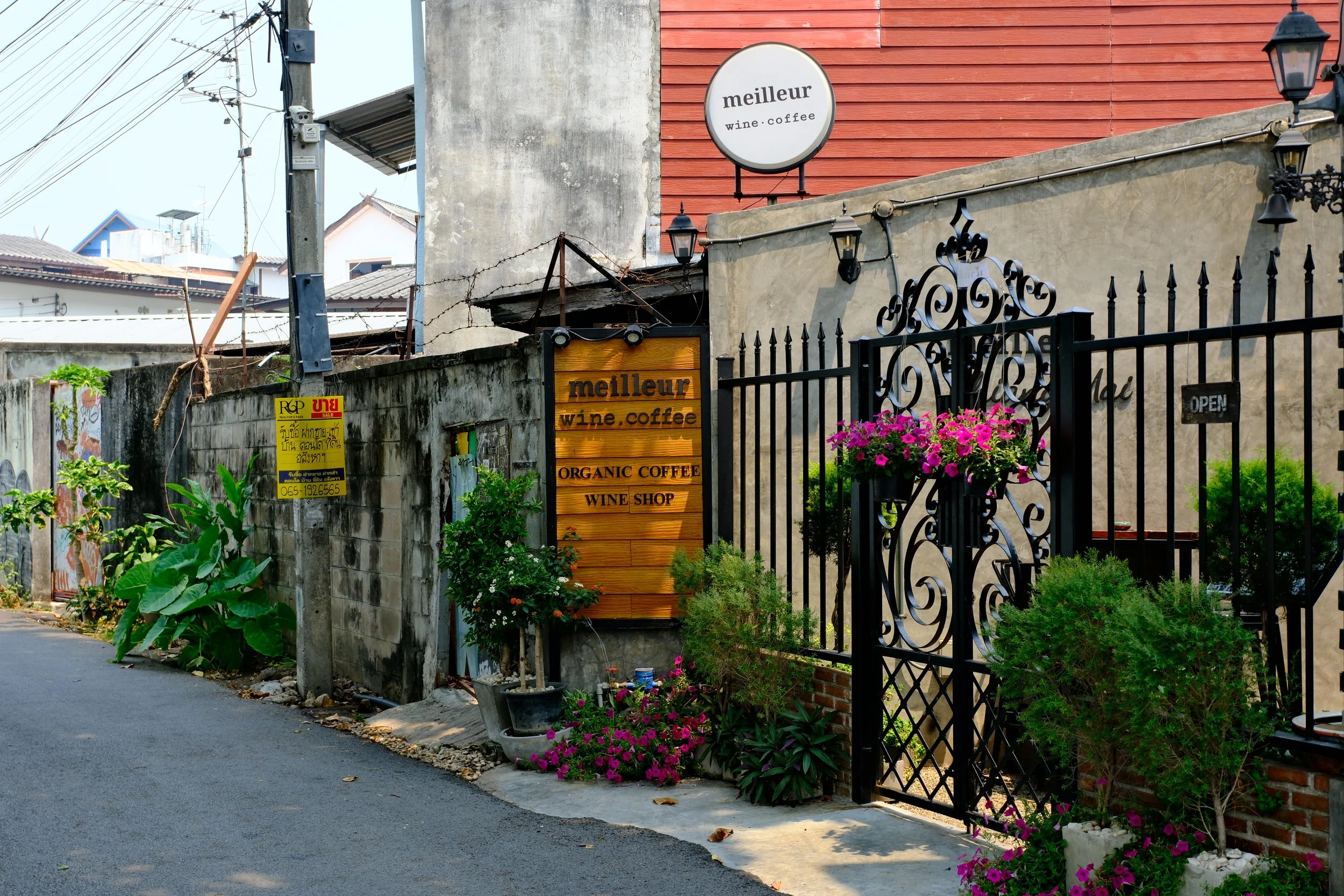 Exterior of a wine and coffee shop named 'meilleur' with decorative black wrought iron gate, pink flowers, and small green bushes. A round signboard with 'meilleur wine coffee' hangs above, and a wooden signboard lists 'Organic Coffee' and 'Wine Shop