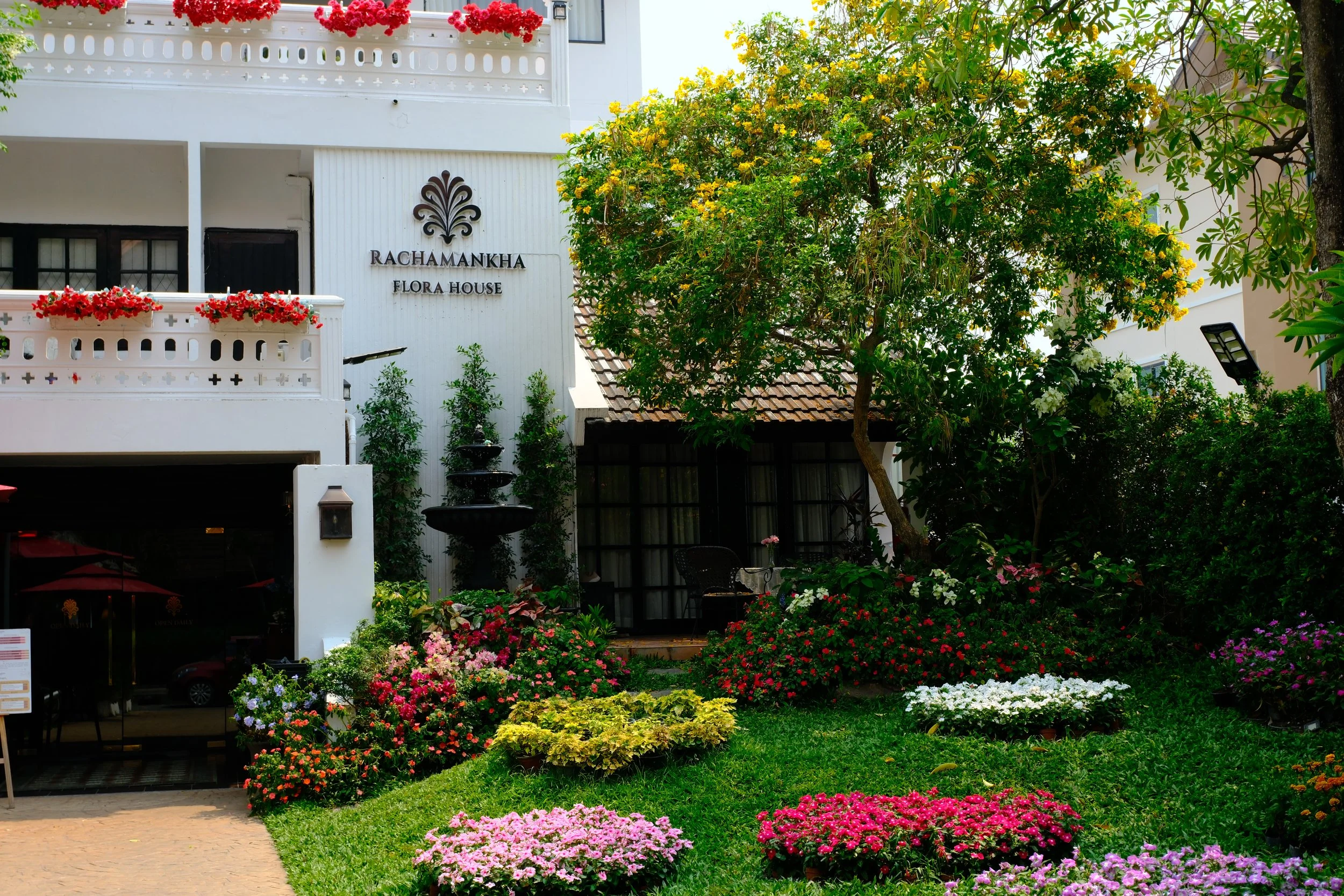 A garden with colorful flowers, a tree, and a white building with balconies and a sign that reads RACHAMANKHA FLORA HOUSE.