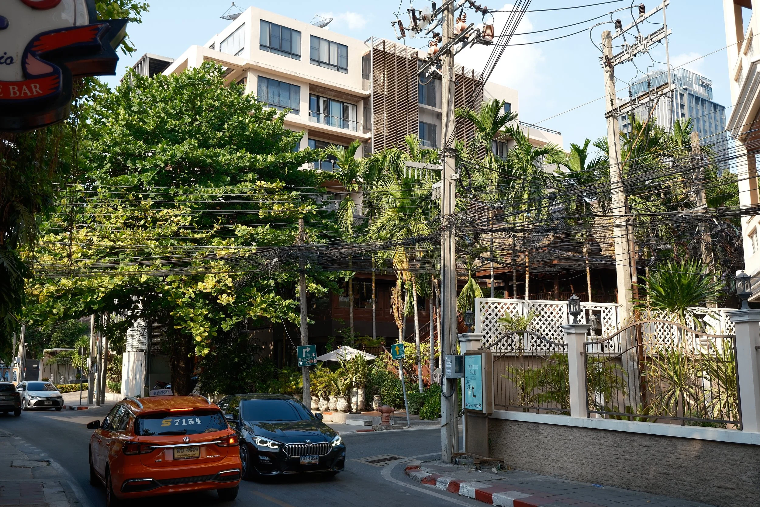 A street scene with cars parked and driving, lush green trees, power lines, and modern apartment buildings in the background.
