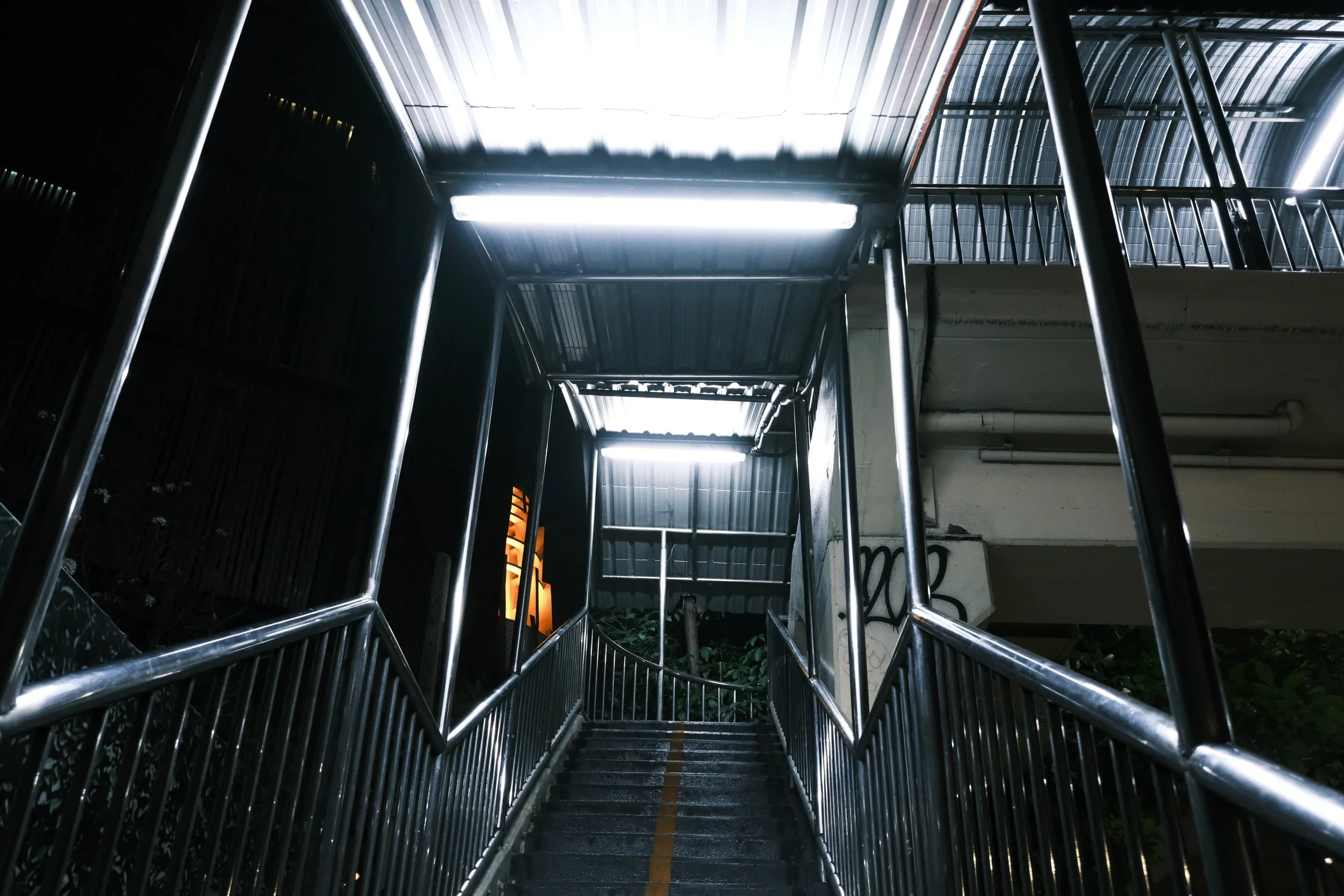 Outdoor metal staircase with illuminated overhead lights and a yellow line down the center, leading up to a walkway under a metal roof.