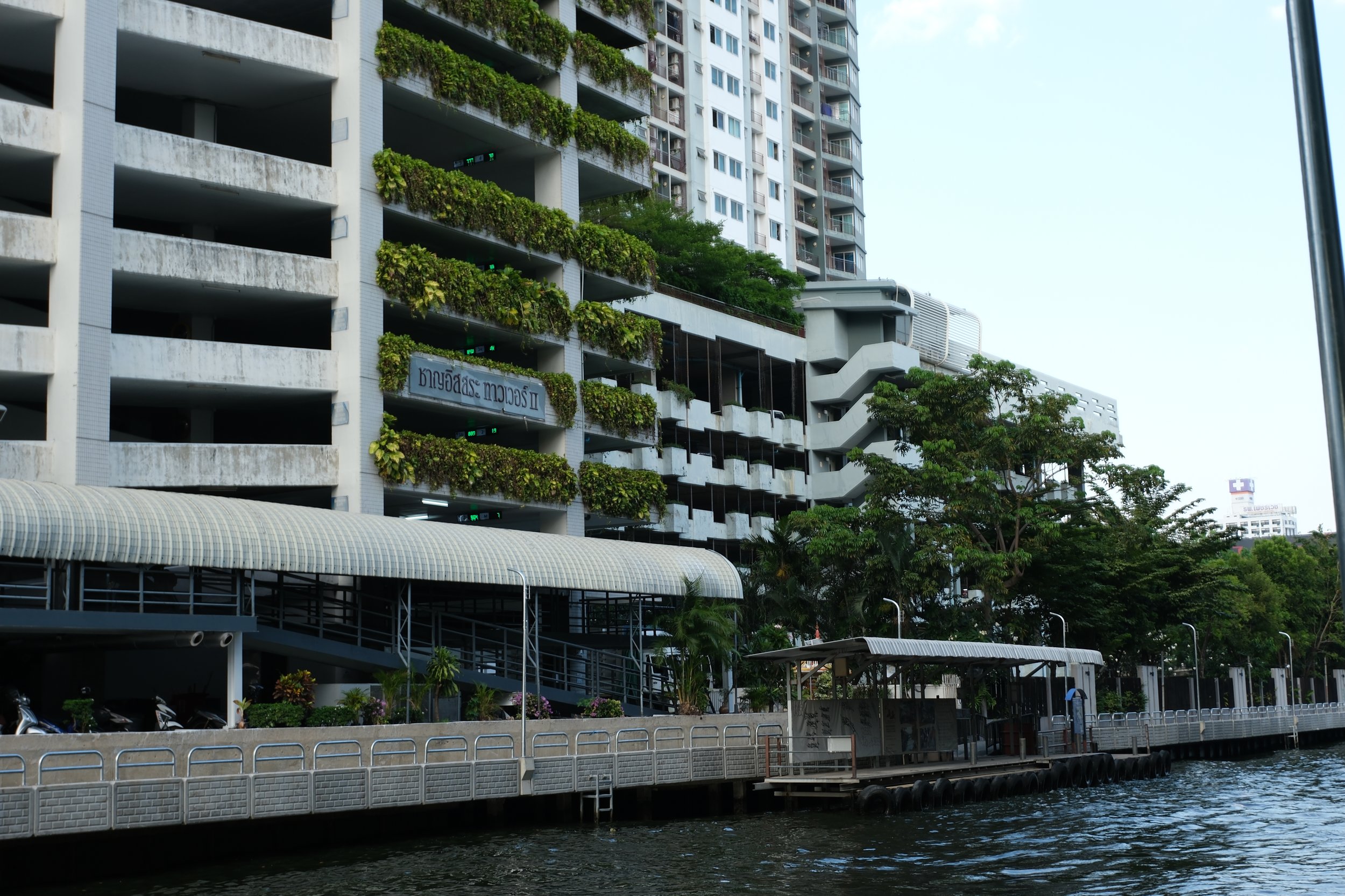 A large modern building situated along a waterway, with balconies filled with green plants, trees lining the water's edge, and a covered walkway along the water.