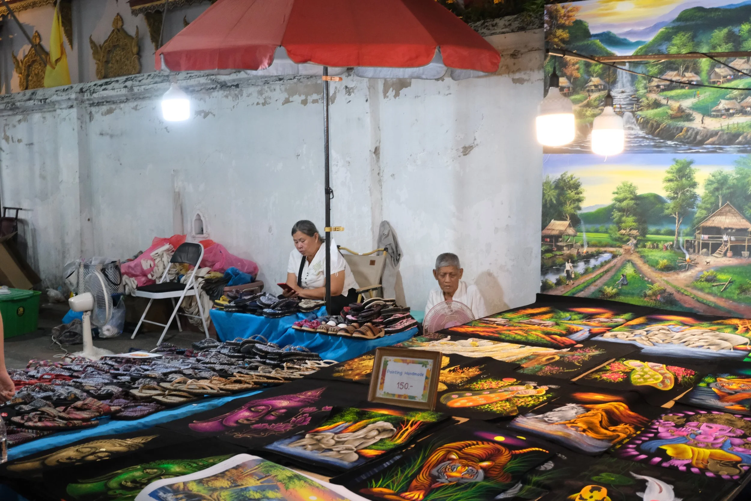 Street market stall selling painted handbags with vibrant jungle and animal designs, with two vendors behind the display and a large landscape mural in the background.