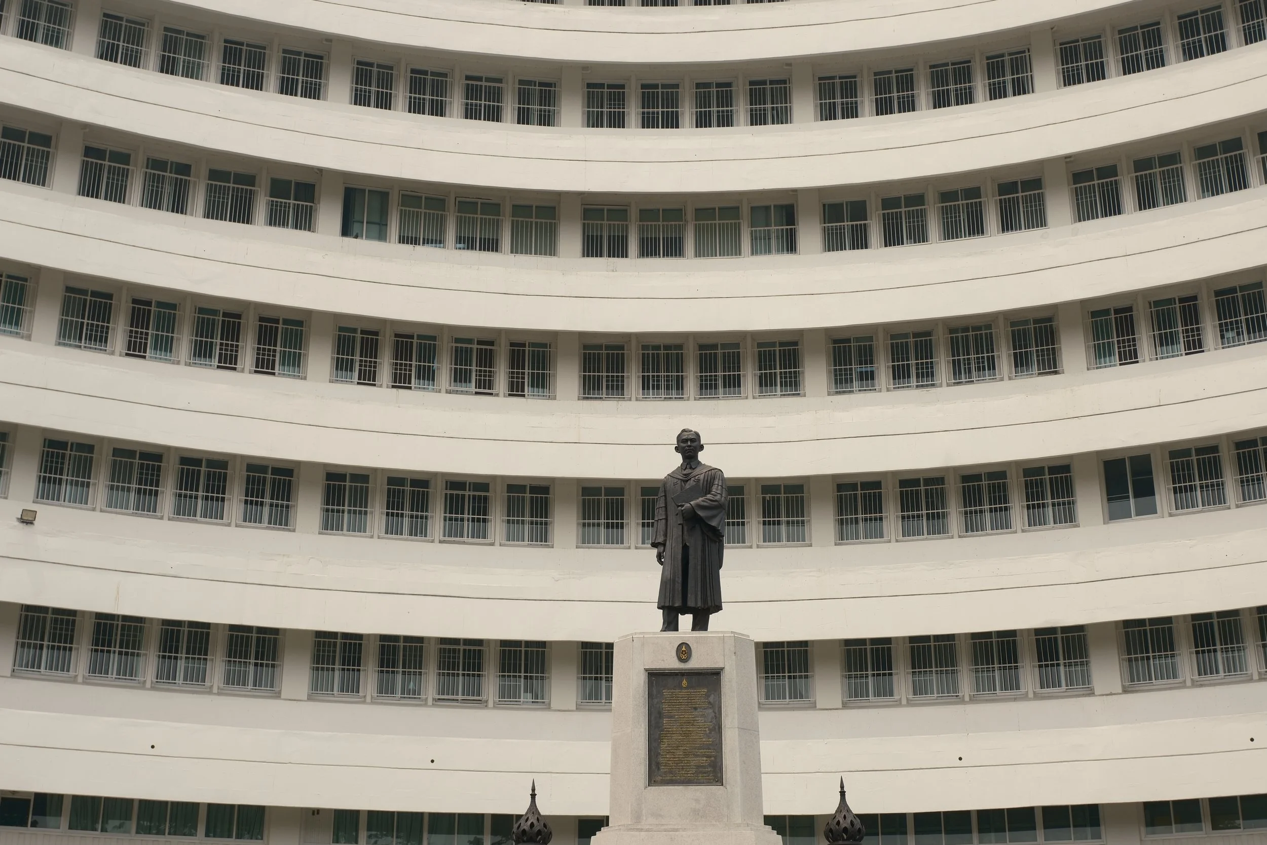 Statue of a man in academic robes on a pedestal in front of a curved white building with multiple floors and repeating window patterns.