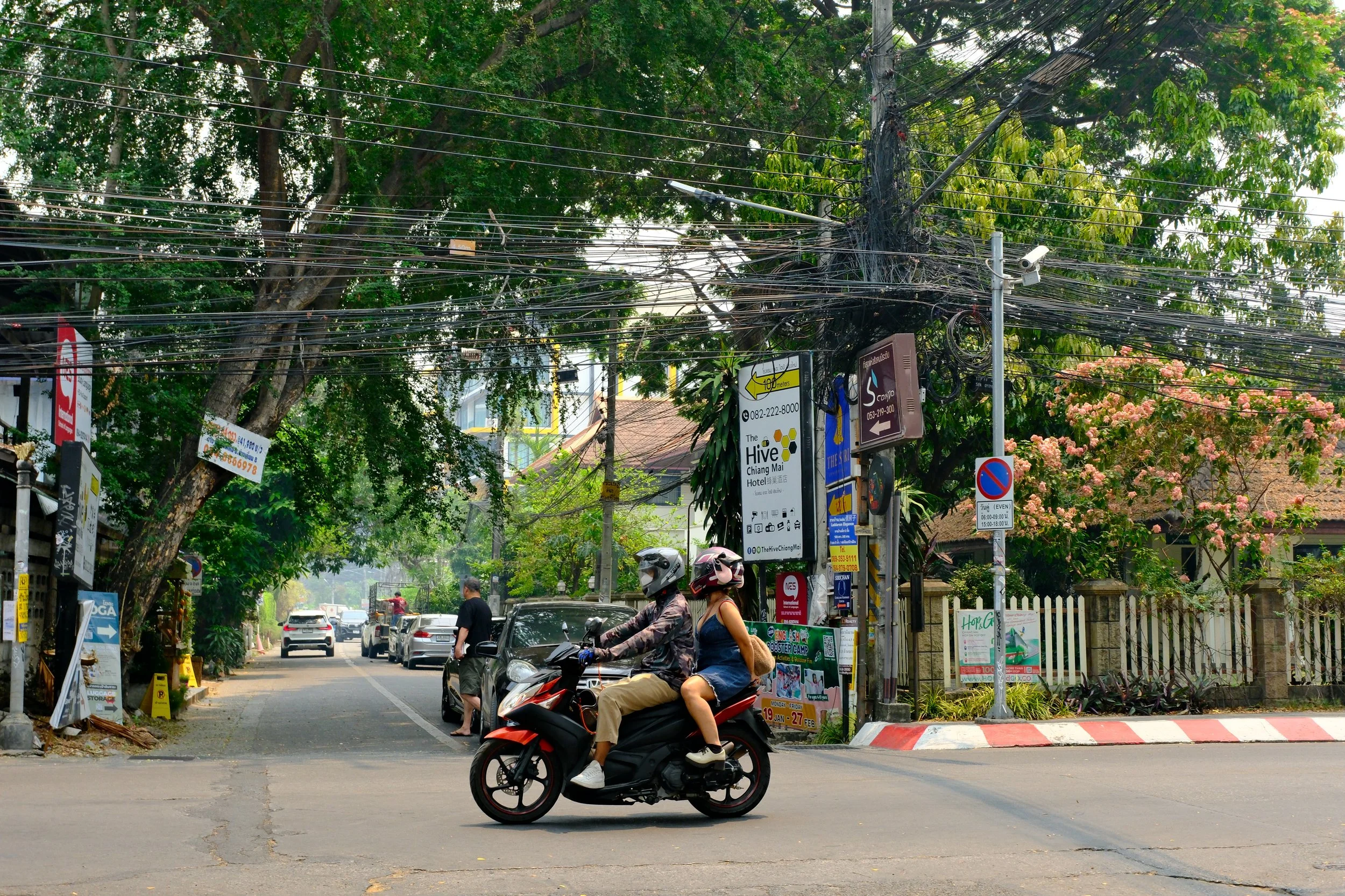 Two people riding a red and black scooter on a street with trees and parked cars, and a man walking nearby in a city with signs and power lines.
