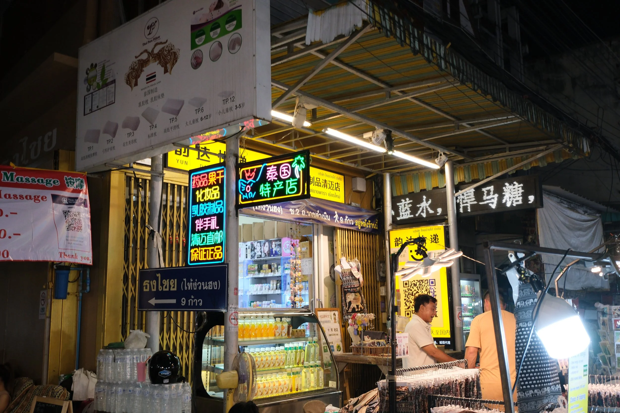 Night scene of a street shop in Thailand with colorful neon signs, displayed products, and two people conversing, under a striped awning.