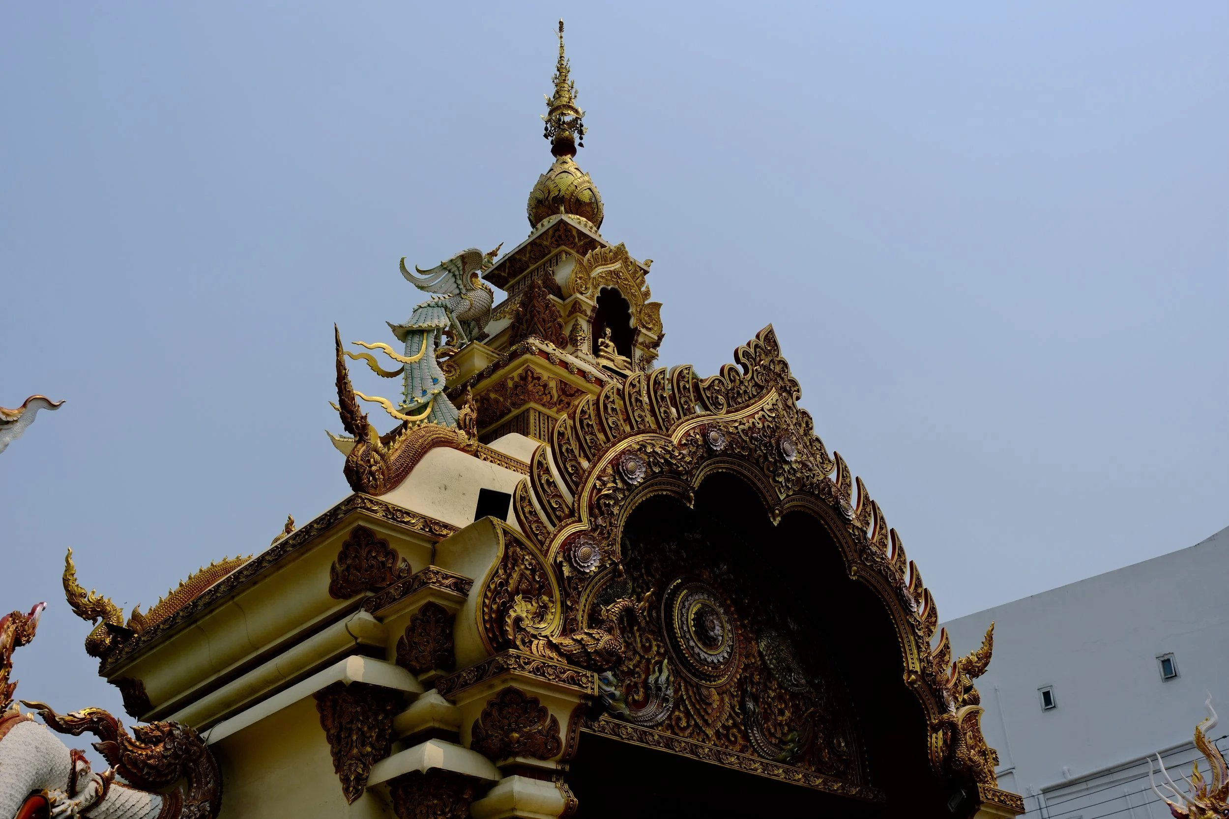 Close-up of an ornate Thai temple roof with intricate gold details, dragon and bird sculptures, and a decorative spire against a clear blue sky.