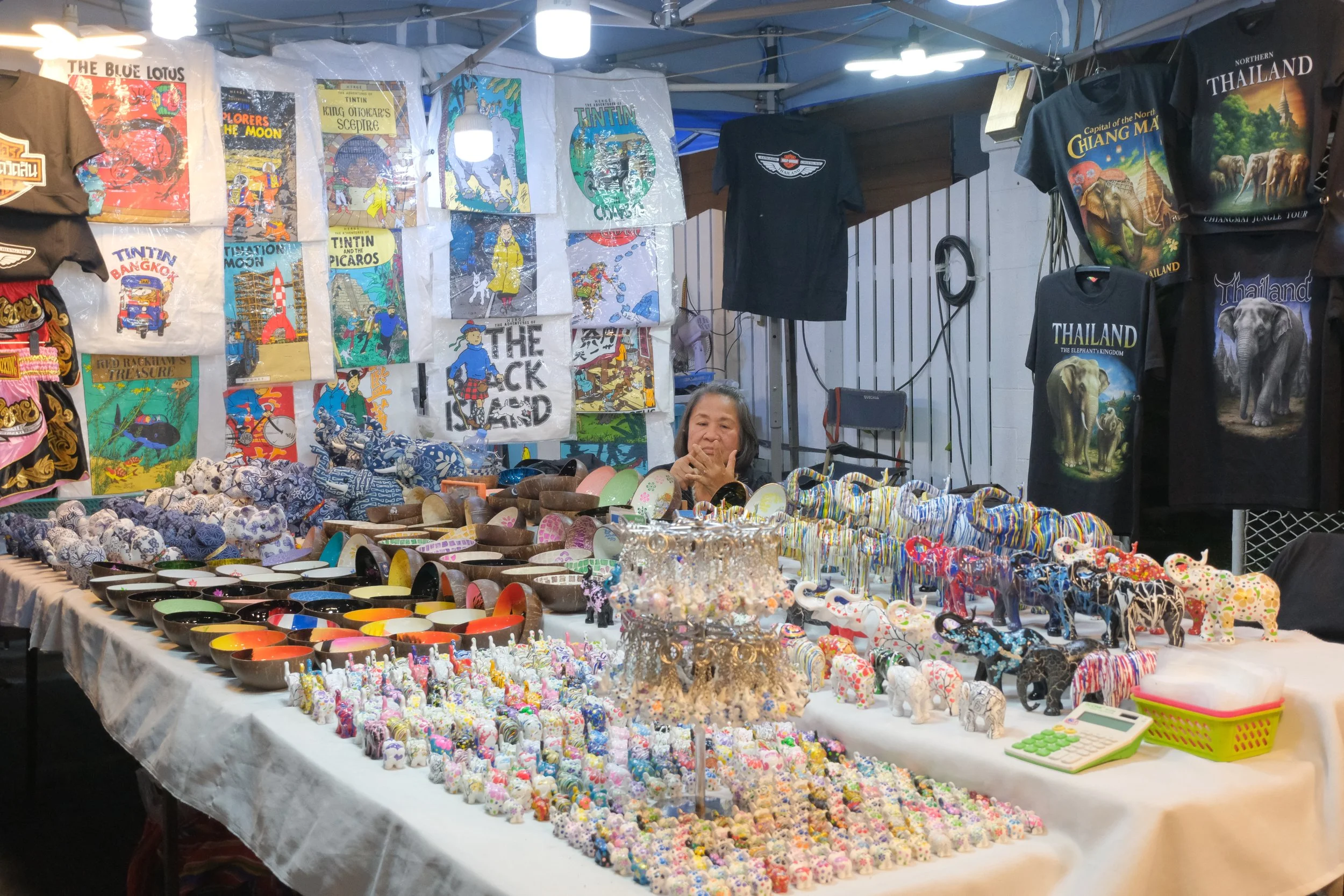 A street vendor stall displays colorful jewelry, ceramic elephants, and souvenirs with a woman behind the table. T-shirts with elephant and Thai tourism designs hang on the background.