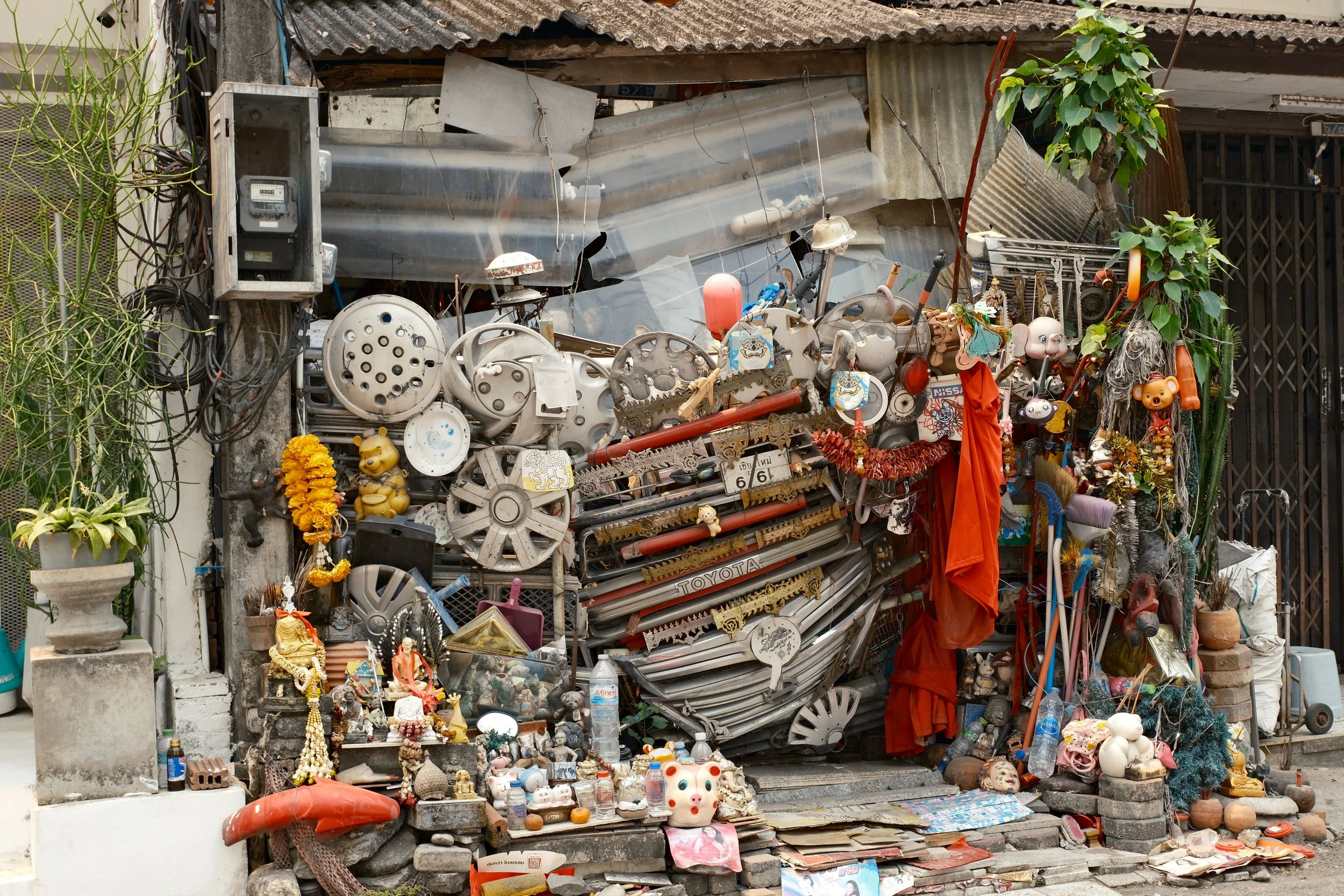 A cluttered outdoor scene with various items including old vehicle parts, toys, statues, plants, and household objects arranged in a haphazard manner.